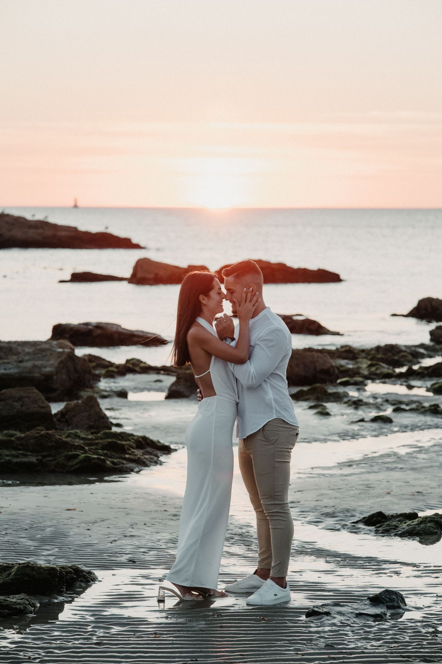 A romantic couple embraces on a rocky beach at sunset with ocean waves and cliffs in the background.