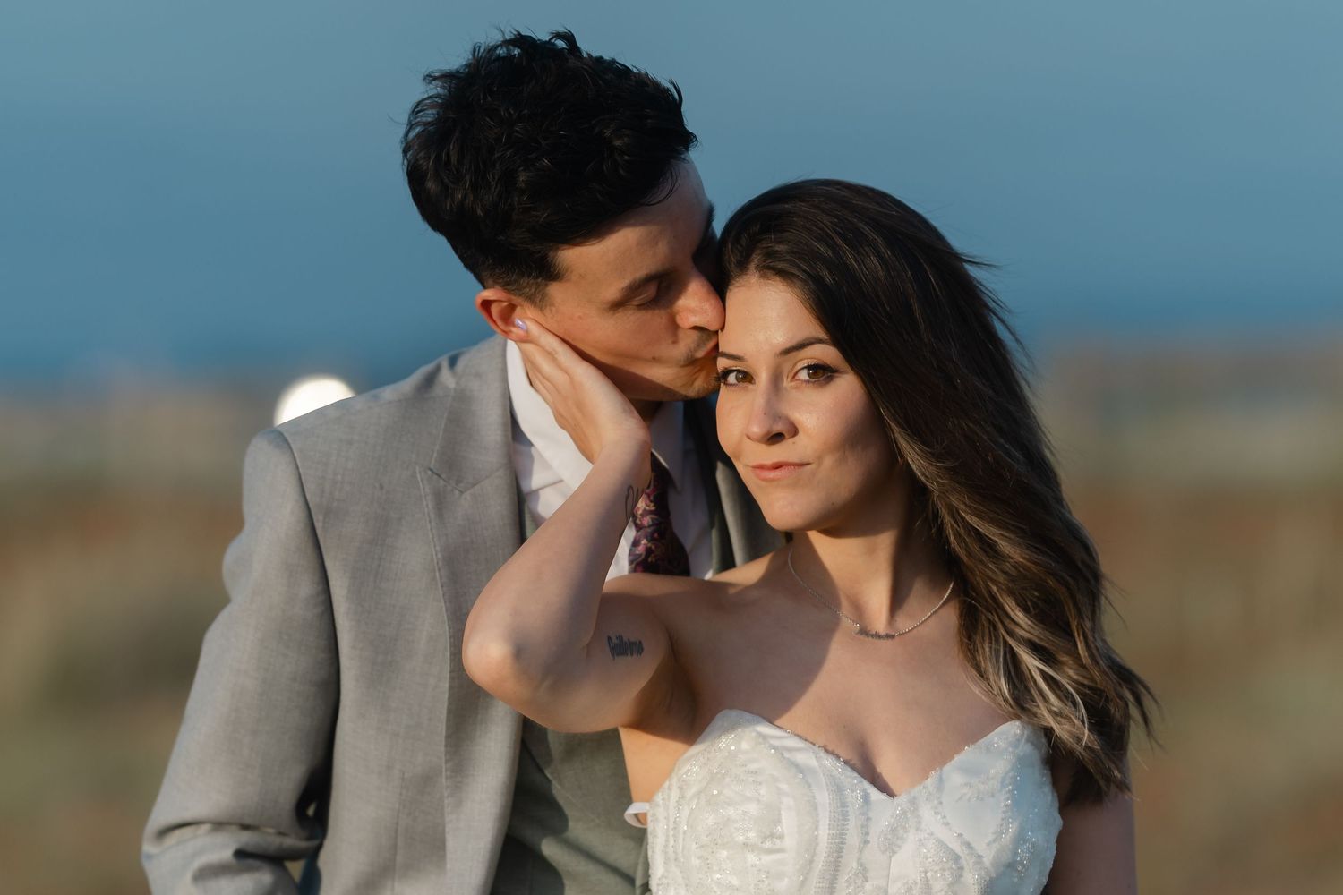 A groom in a gray suit tenderly kisses his bride's temple as she smiles softly in a white strapless wedding gown.