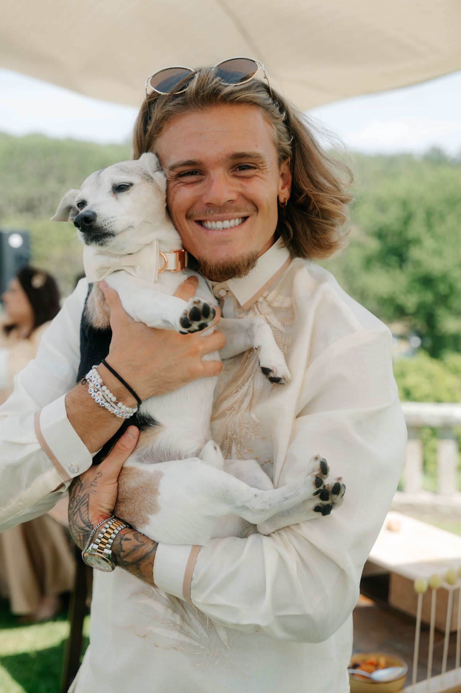 A person in a white outfit holds a small white dog while smiling outdoors on a sunny day.