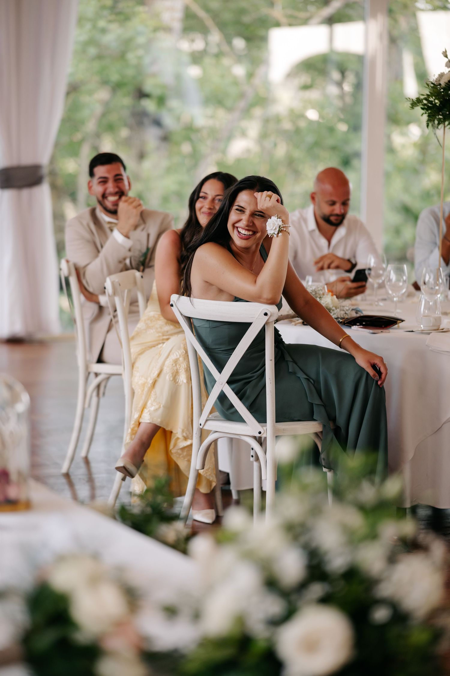Wedding reception guests share joyful laughter at an elegant white-draped venue with floral centerpieces.