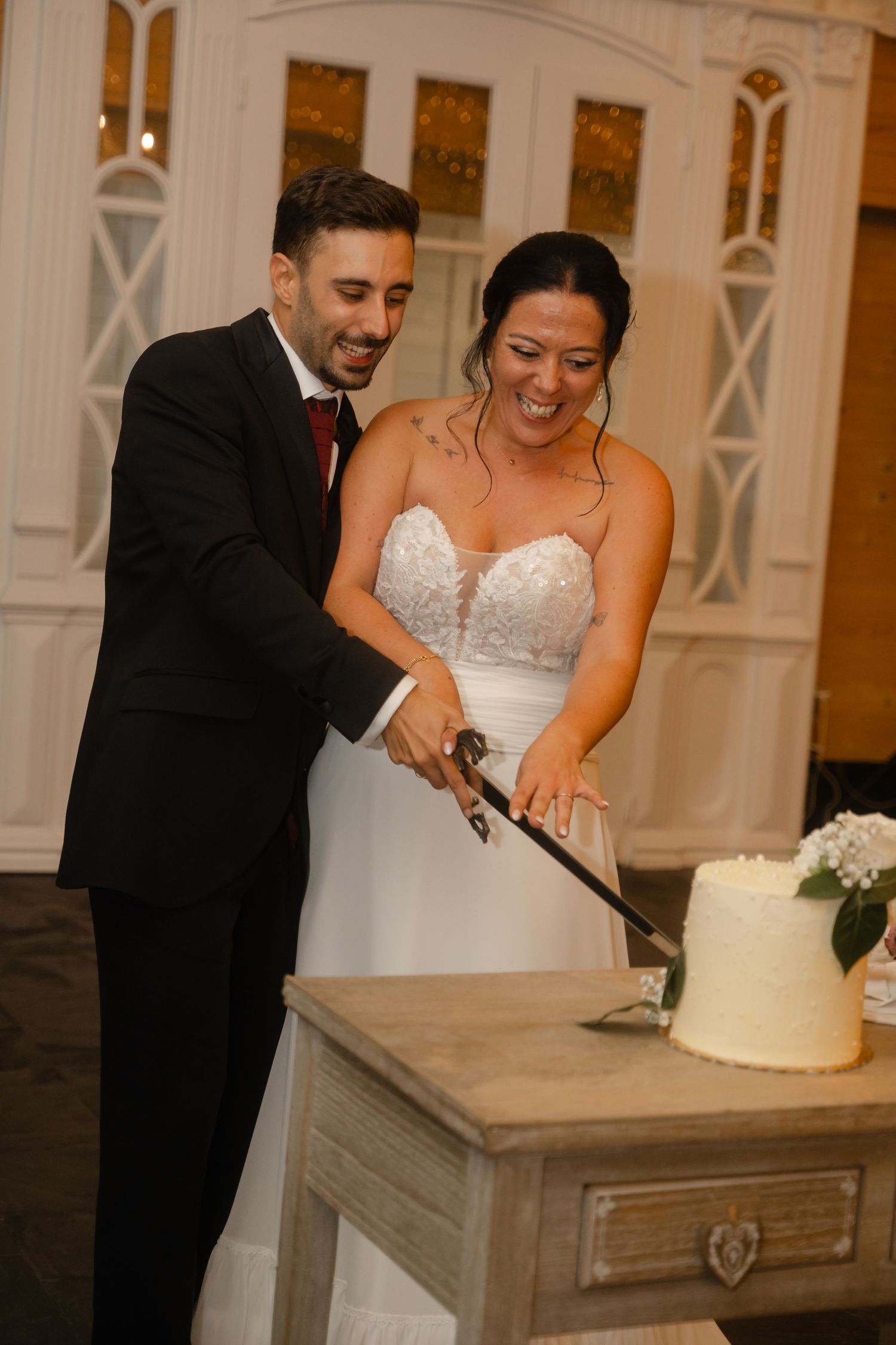 Newlywed couple cuts their elegant white wedding cake together at an indoor reception venue.
