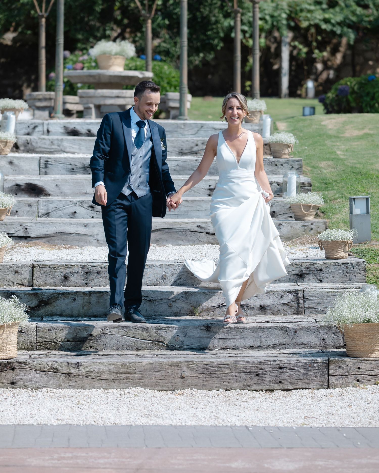 A couple in wedding attire walking together across stone steps in an outdoor garden setting.