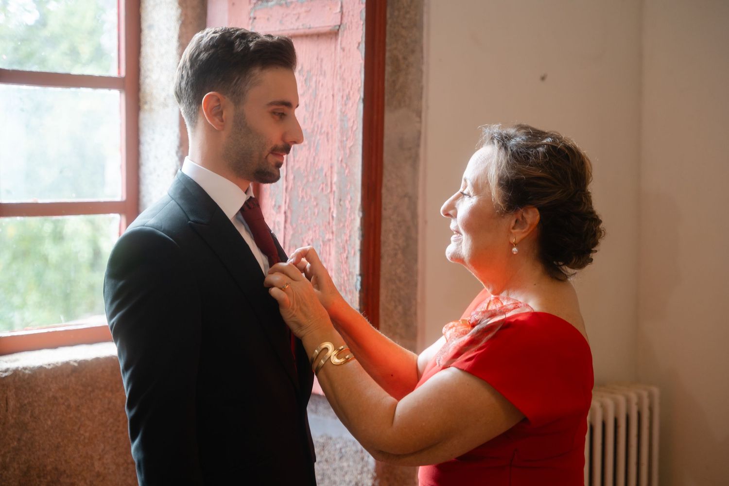 A tender moment between two people as one adjusts the other's tie in a room with red walls and natural light.