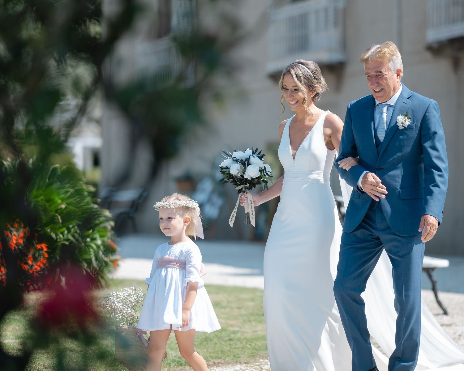 A couple in wedding attire walks through a garden setting while a young child in white runs ahead.