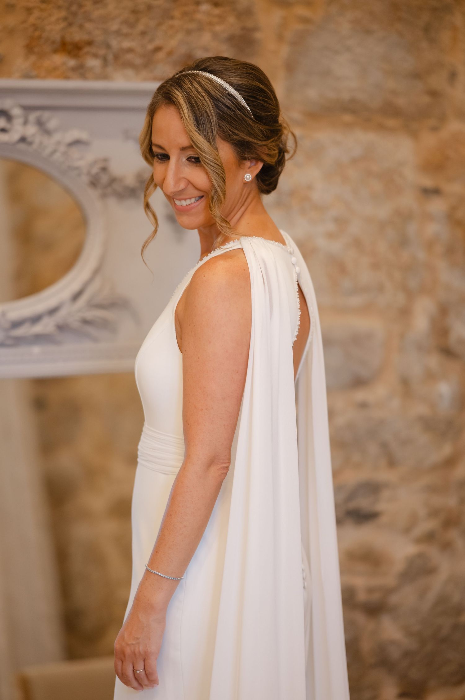 Elegant bride in a flowing white wedding gown with cape detail poses against a textured stone wall.