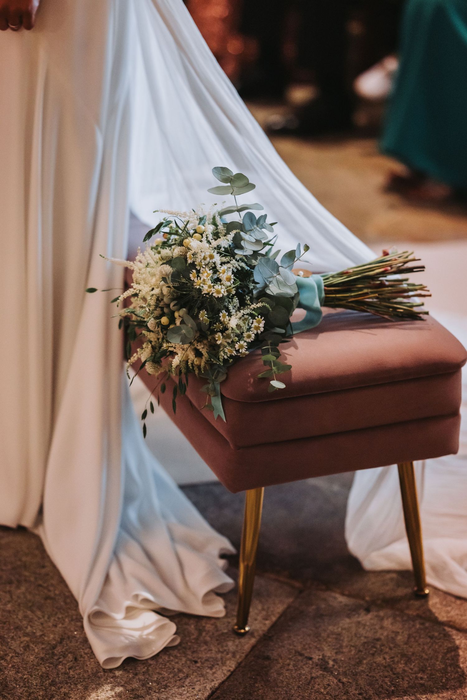 A white floral wedding bouquet rests on a wooden pew in a church with draped white fabric.