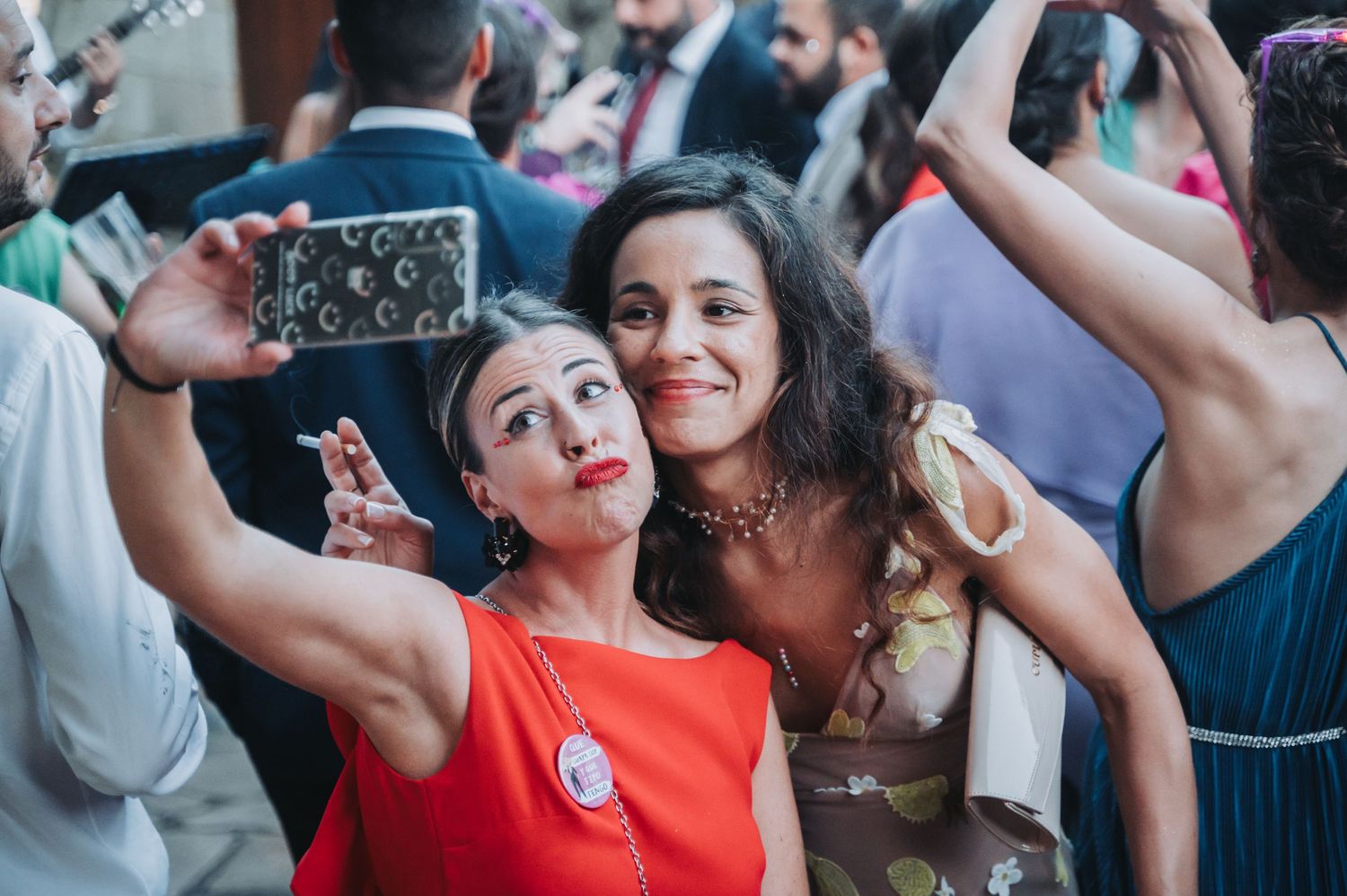 Two people take a fun selfie together at a crowded event while making silly faces and gestures.