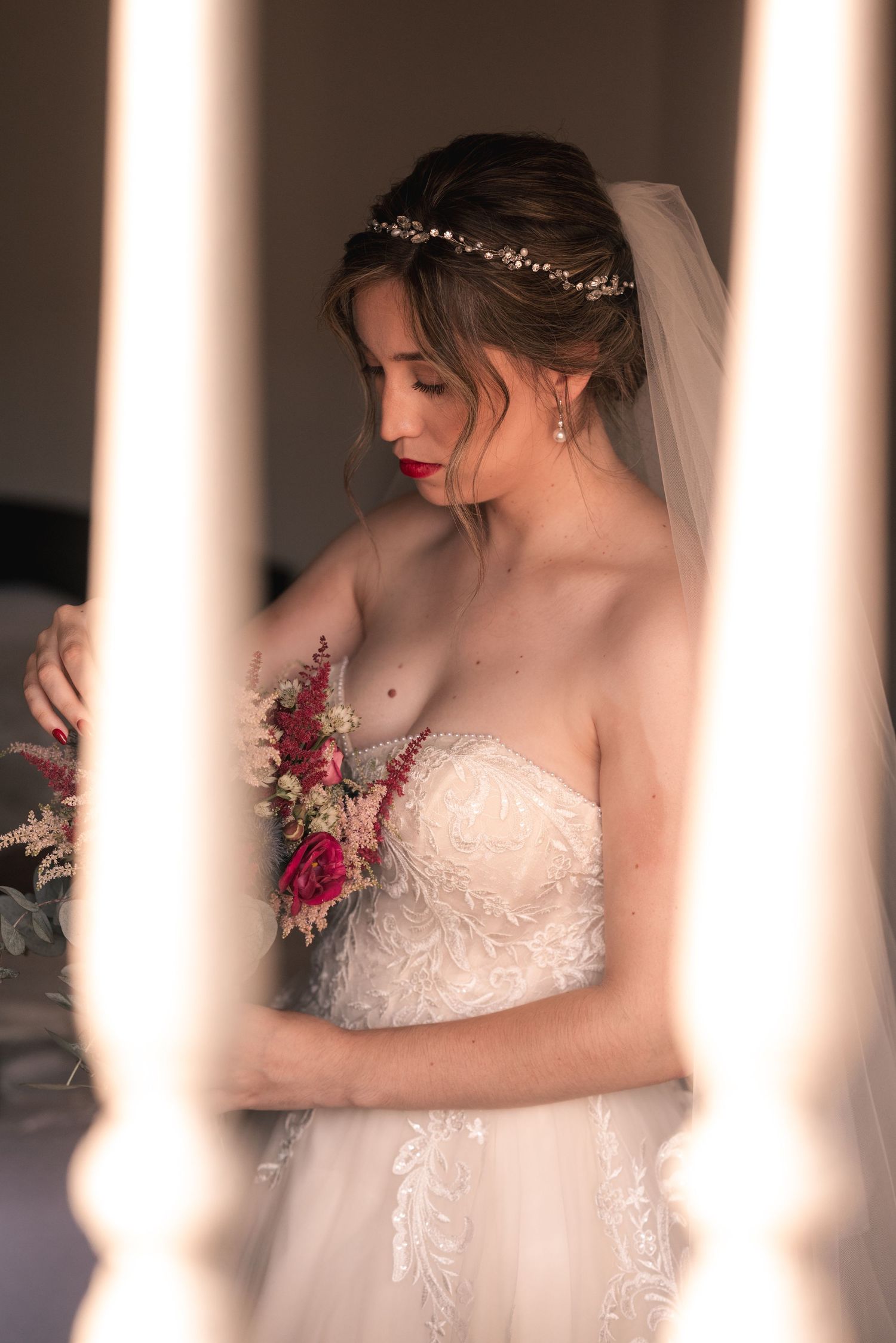 An elegant bridal portrait showing a strapless lace gown, veil and red bouquet through white columns.