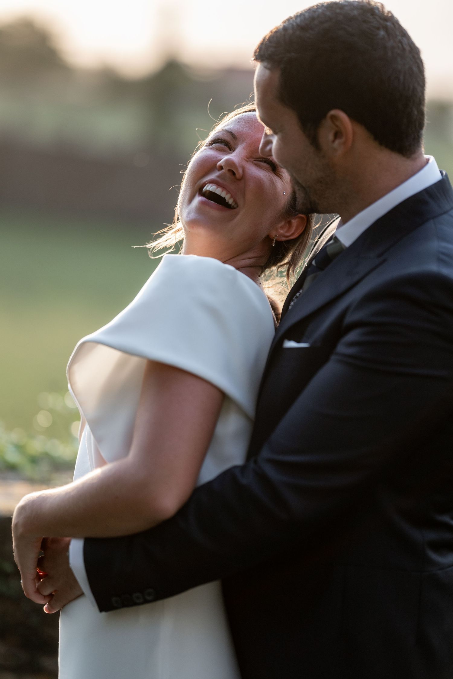 A romantic wedding couple embraces and laughs joyfully during their sunset photo session in an outdoor setting.