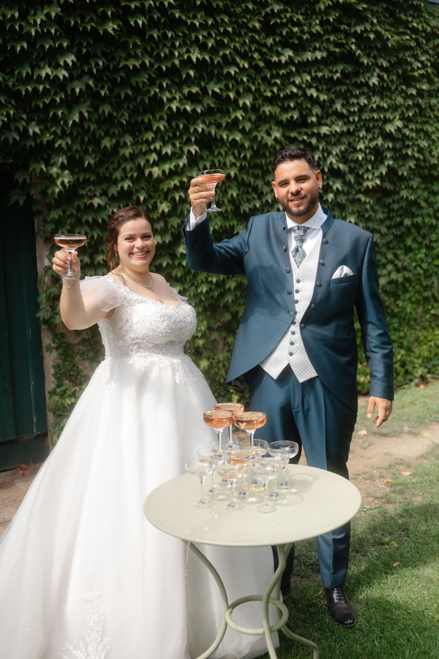 Wedding couple celebrates with champagne glasses at an outdoor garden reception with ivy-covered walls.