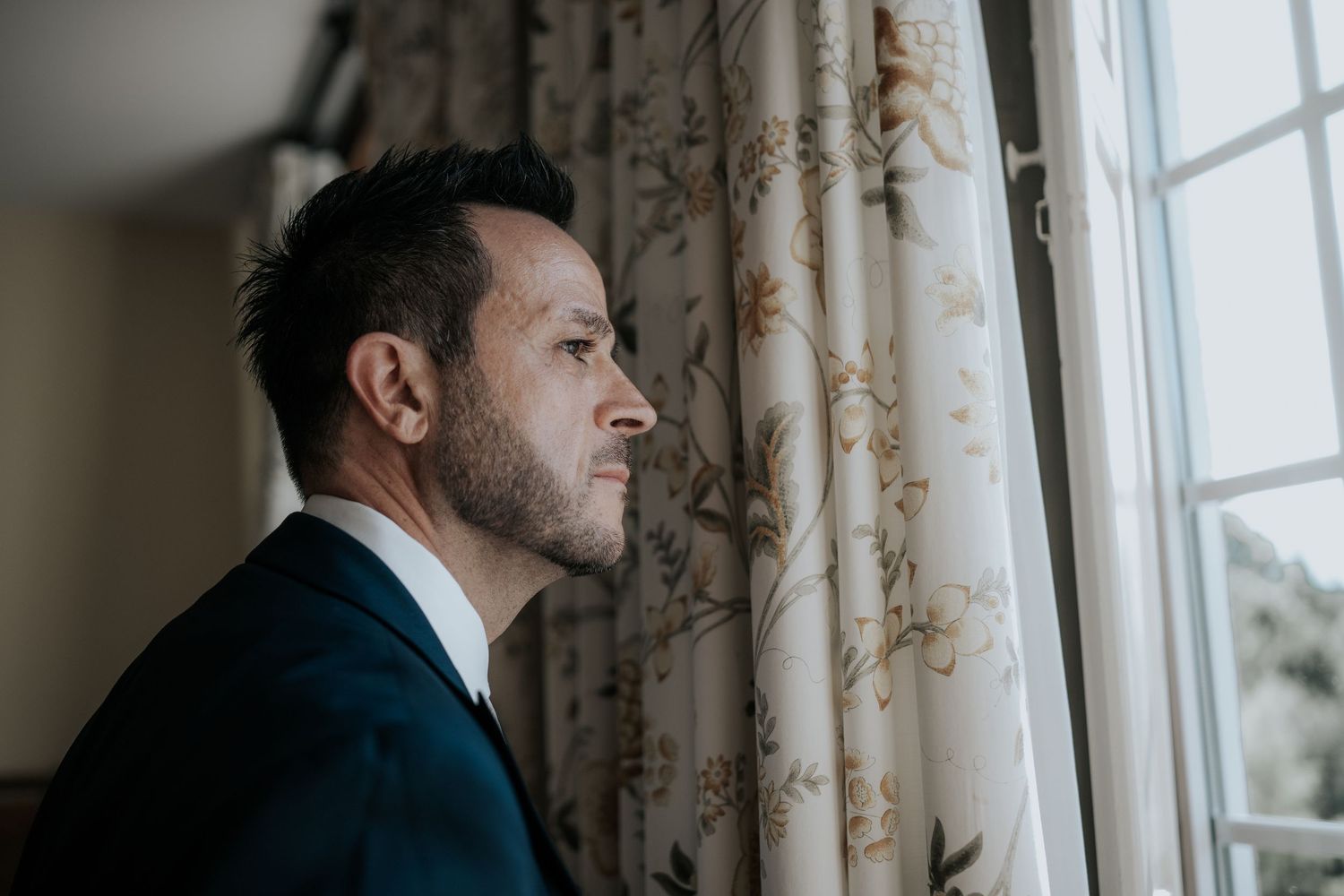 Person in dark suit gazes thoughtfully through window with floral curtains.