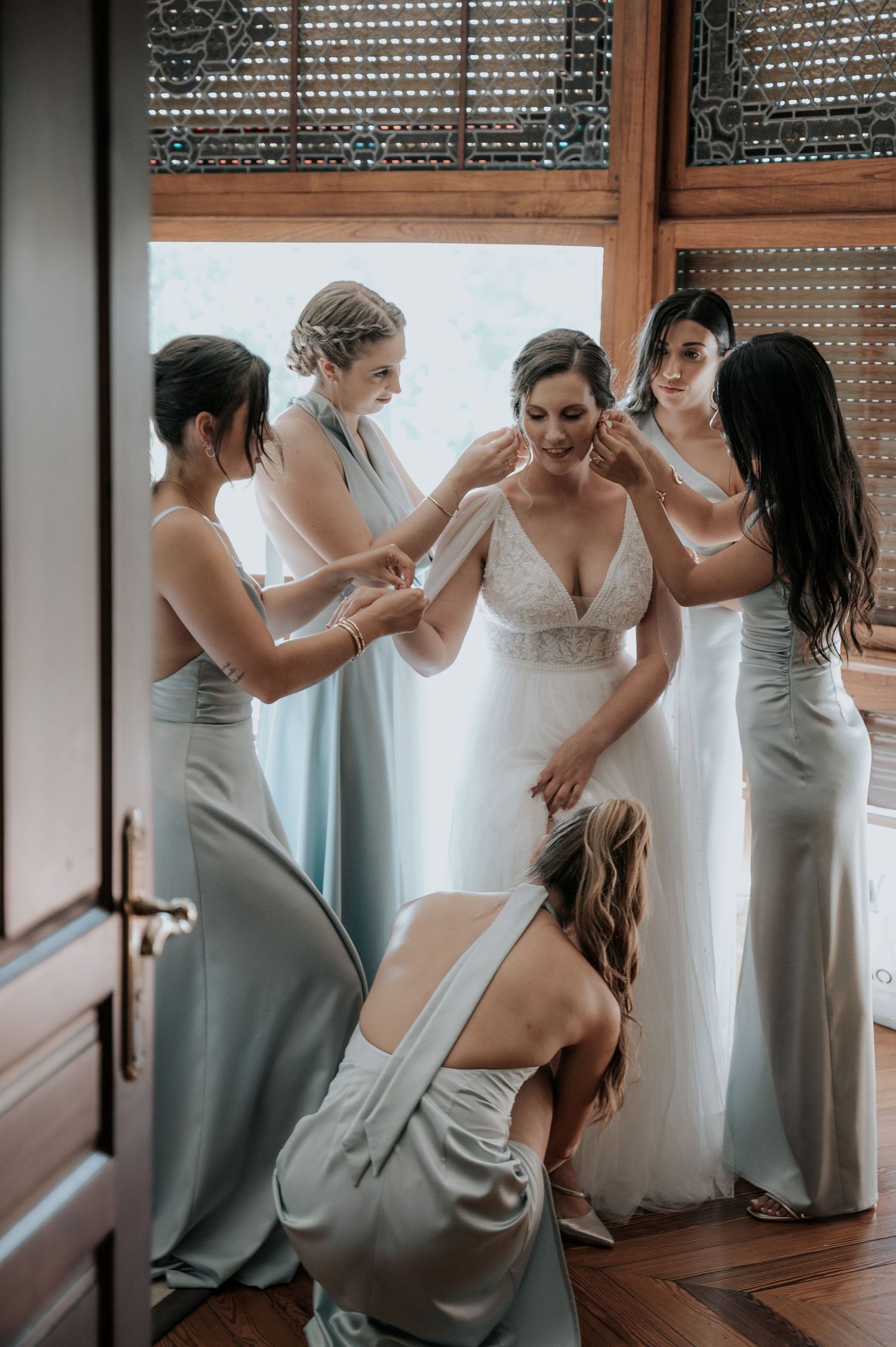 Bridesmaids in sage green dresses help bride get ready in sunlit room.
