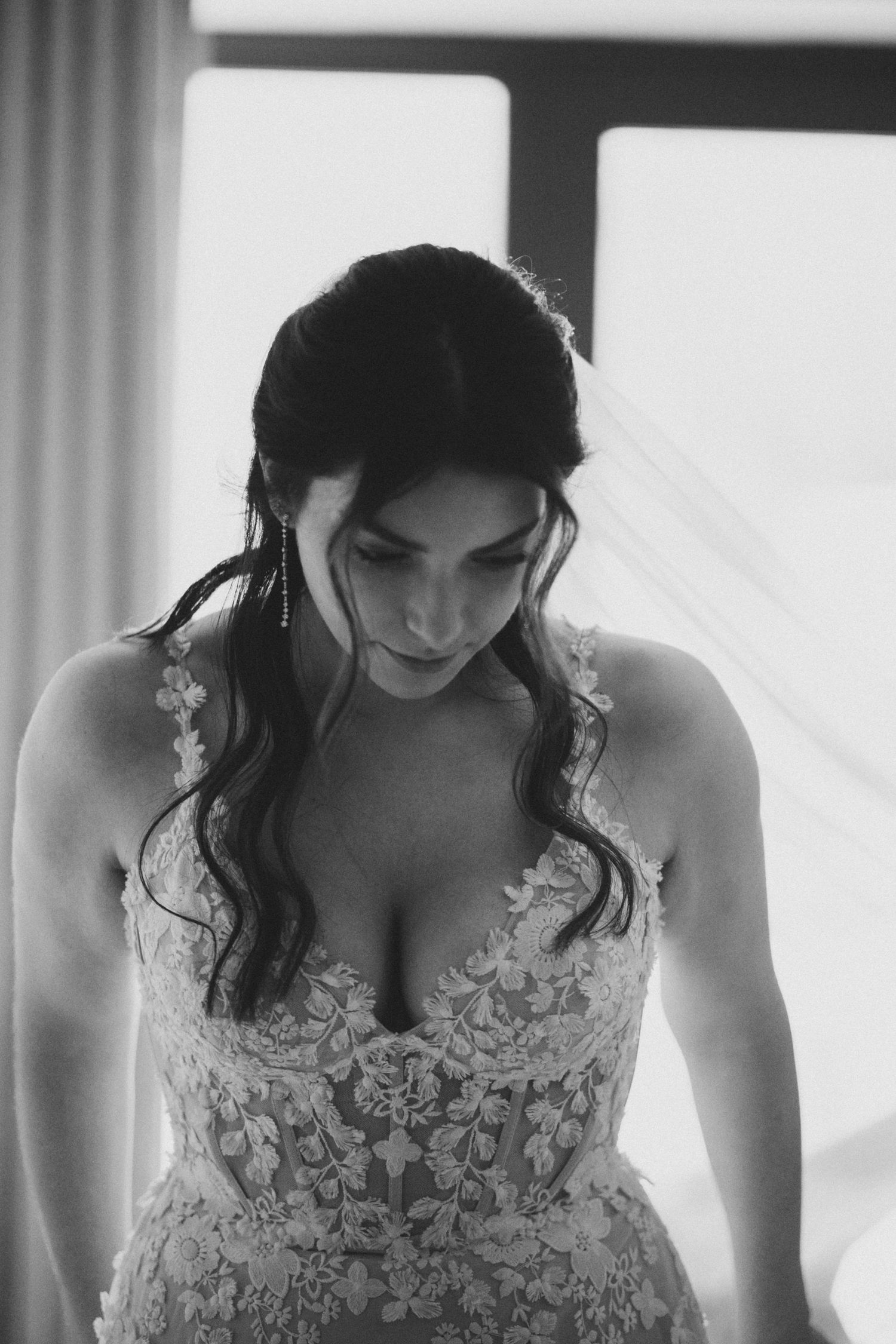 Black and white portrait of a bride looking down, wearing an intricate lace wedding dress with delicate floral details.