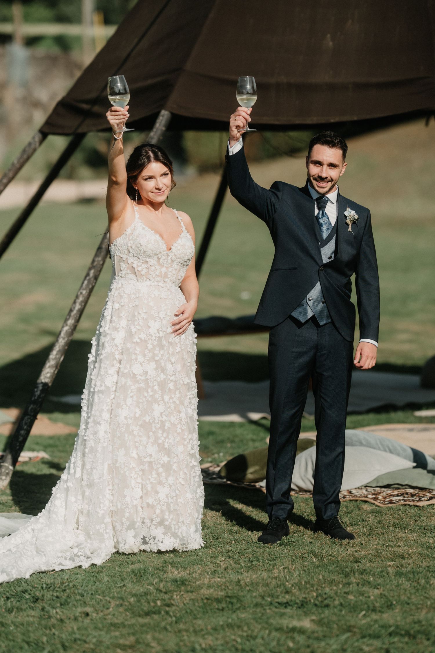 A bride in a lace wedding dress and groom in black suit toast with champagne glasses on a grassy lawn.