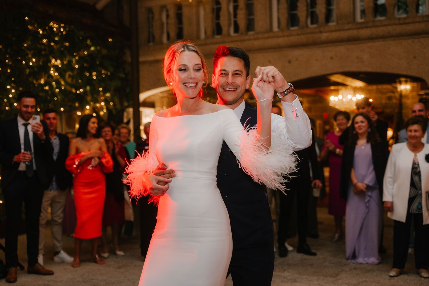 Wedding party celebrates on dance floor with colorful lighting and guests watching in background.