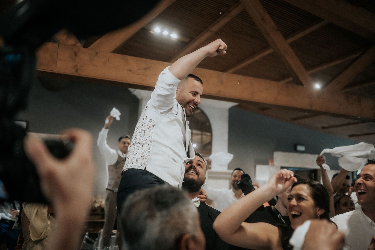 Wedding guests dancing and celebrating on a rustic wooden dance floor with exposed beam ceiling.