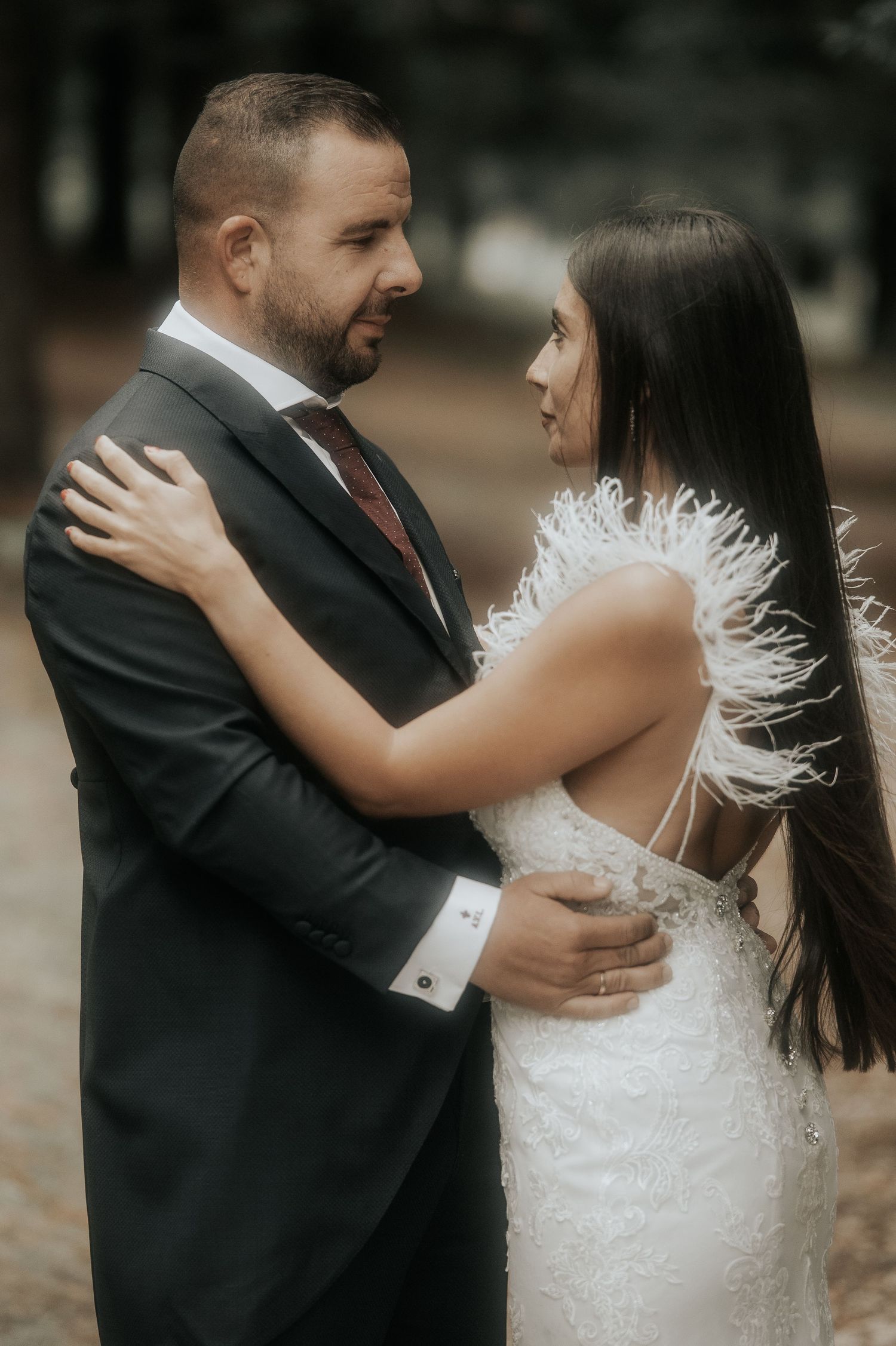 A couple embraces during their wedding, with the bride wearing a feathered white dress and groom in dark suit.