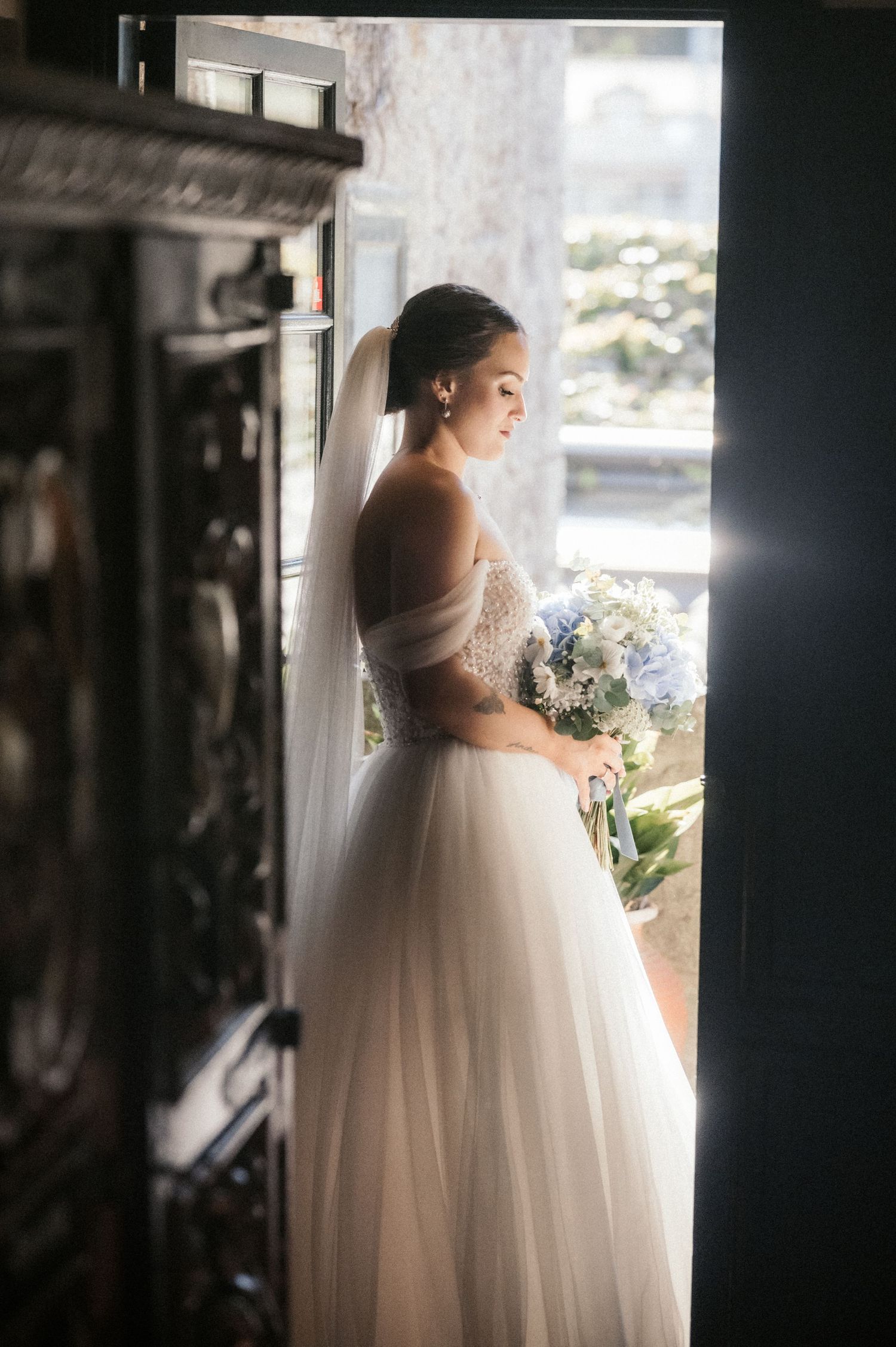 A bride in a flowing white gown stands by a window in silhouette, creating a romantic and ethereal wedding portrait.