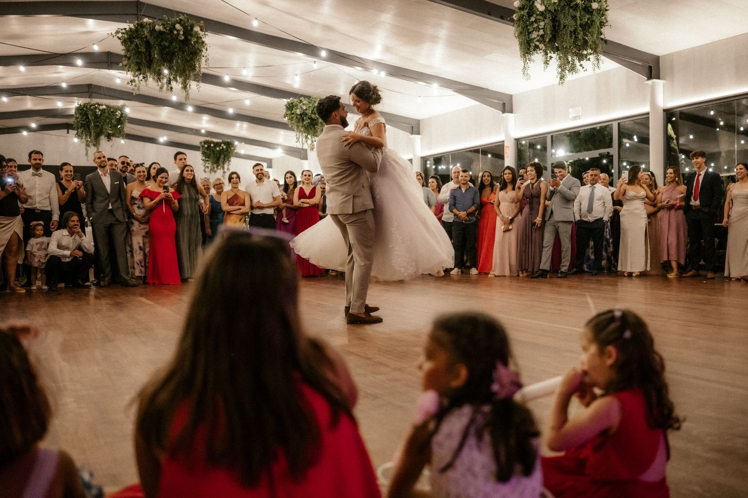 Wedding guests dance together in a spacious reception hall decorated with hanging greenery and string lights.