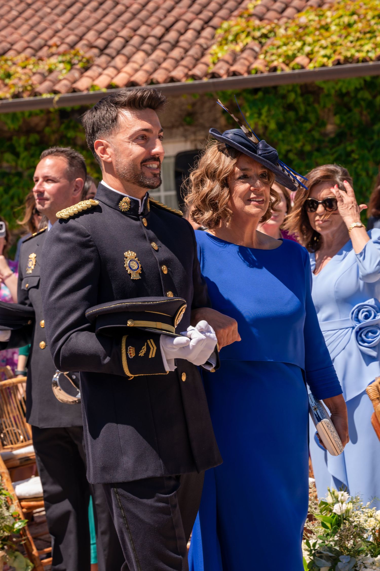 Military officer in dress uniform stands with woman in blue dress and navy hat at formal outdoor event.
