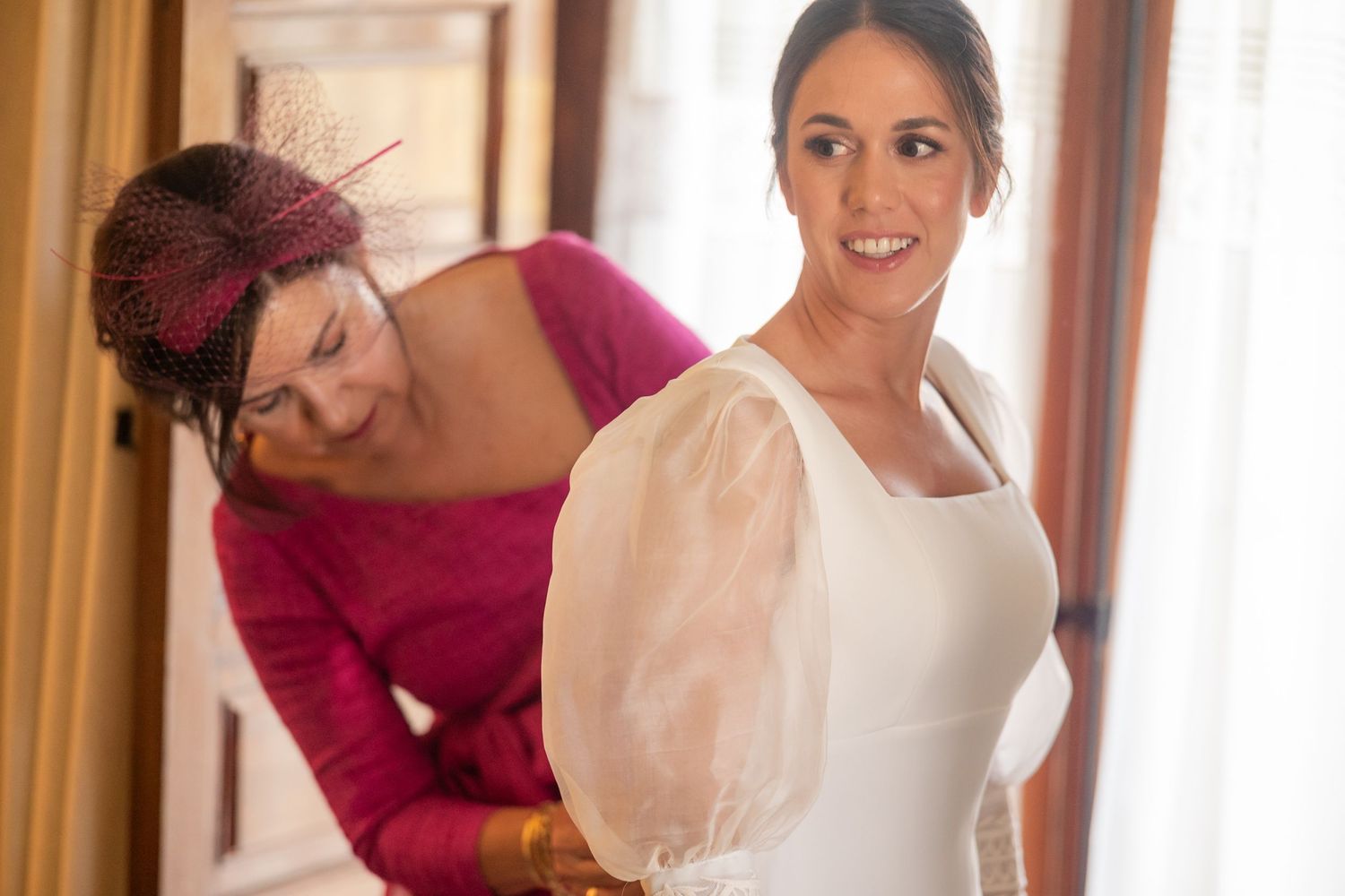 A group of women in formal dresses help adjust a white wedding gown in an elegant indoor setting.