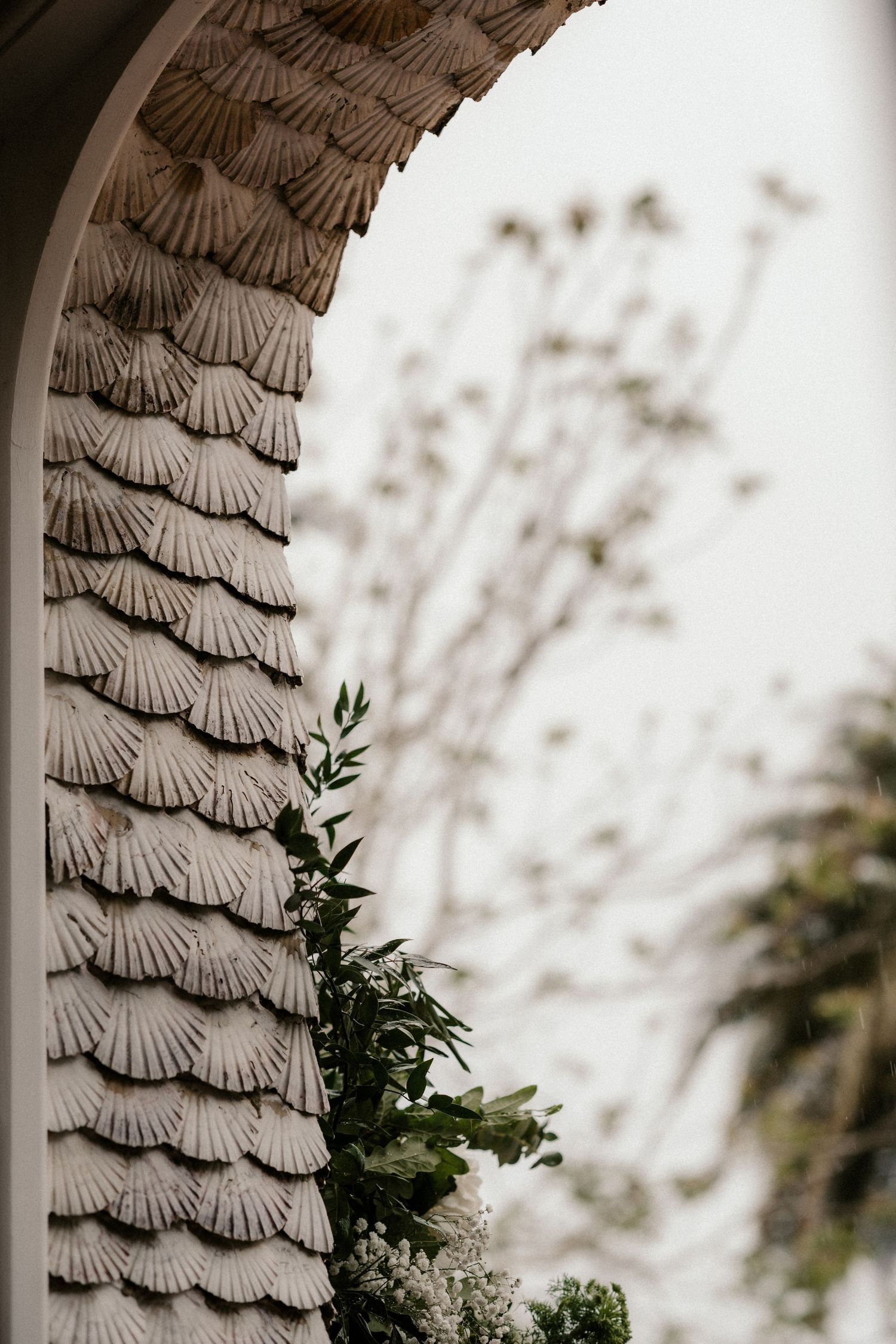Architectural detail showing decorative scalloped shingles on a curved wall with green foliage peeking through.