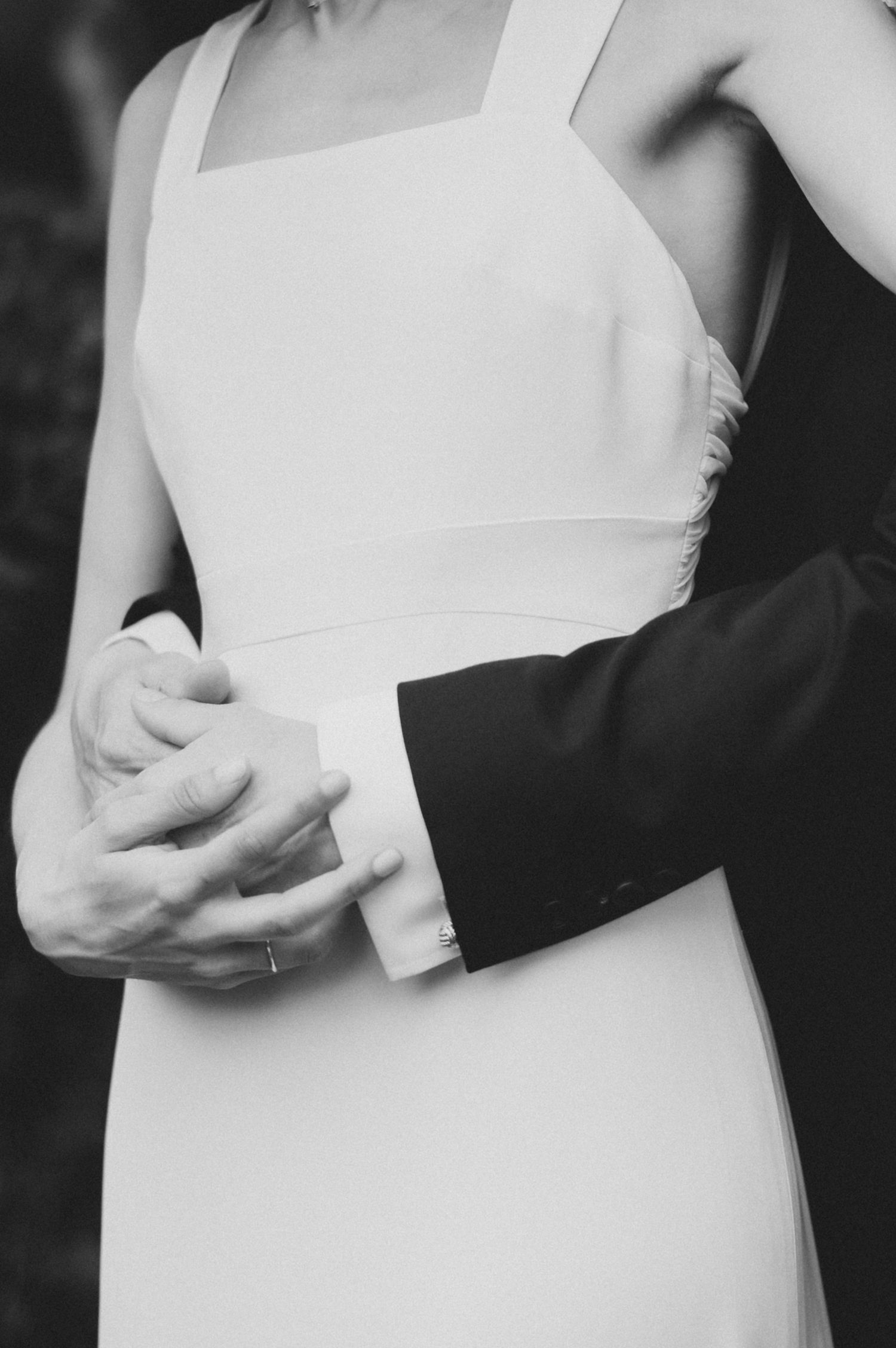 A black and white close-up of clasped hands between a bride in white and groom in dark formal wear.