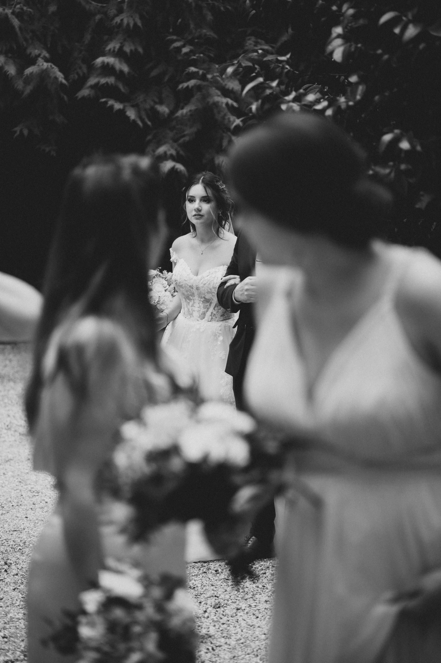 Black and white wedding photo showing a bride in a white dress through blurred foreground figures.