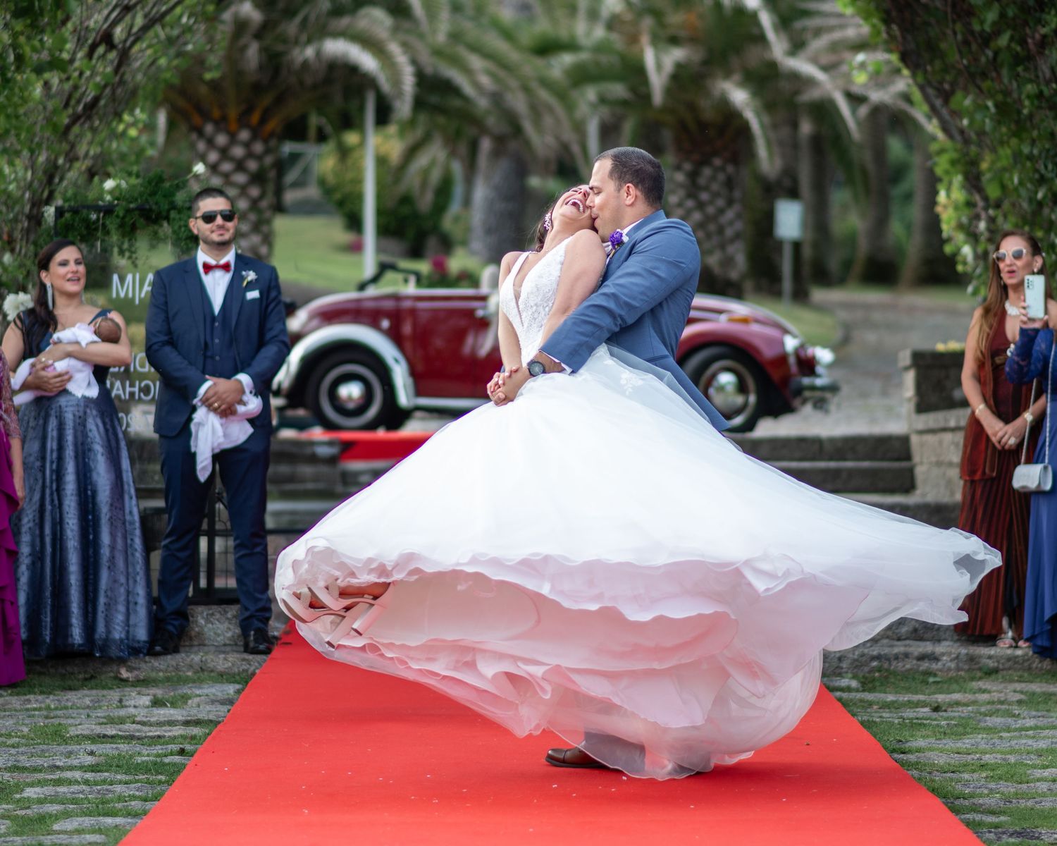 A couple shares a romantic dip during their outdoor wedding dance on a red carpet surrounded by guests.
