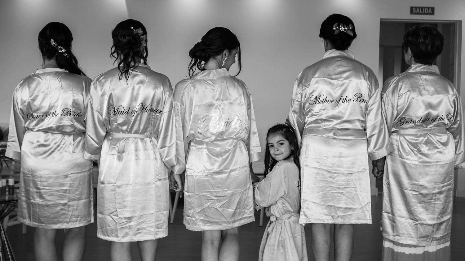 Row of bridesmaids in matching white satin robes with a young flower girl peeking through.