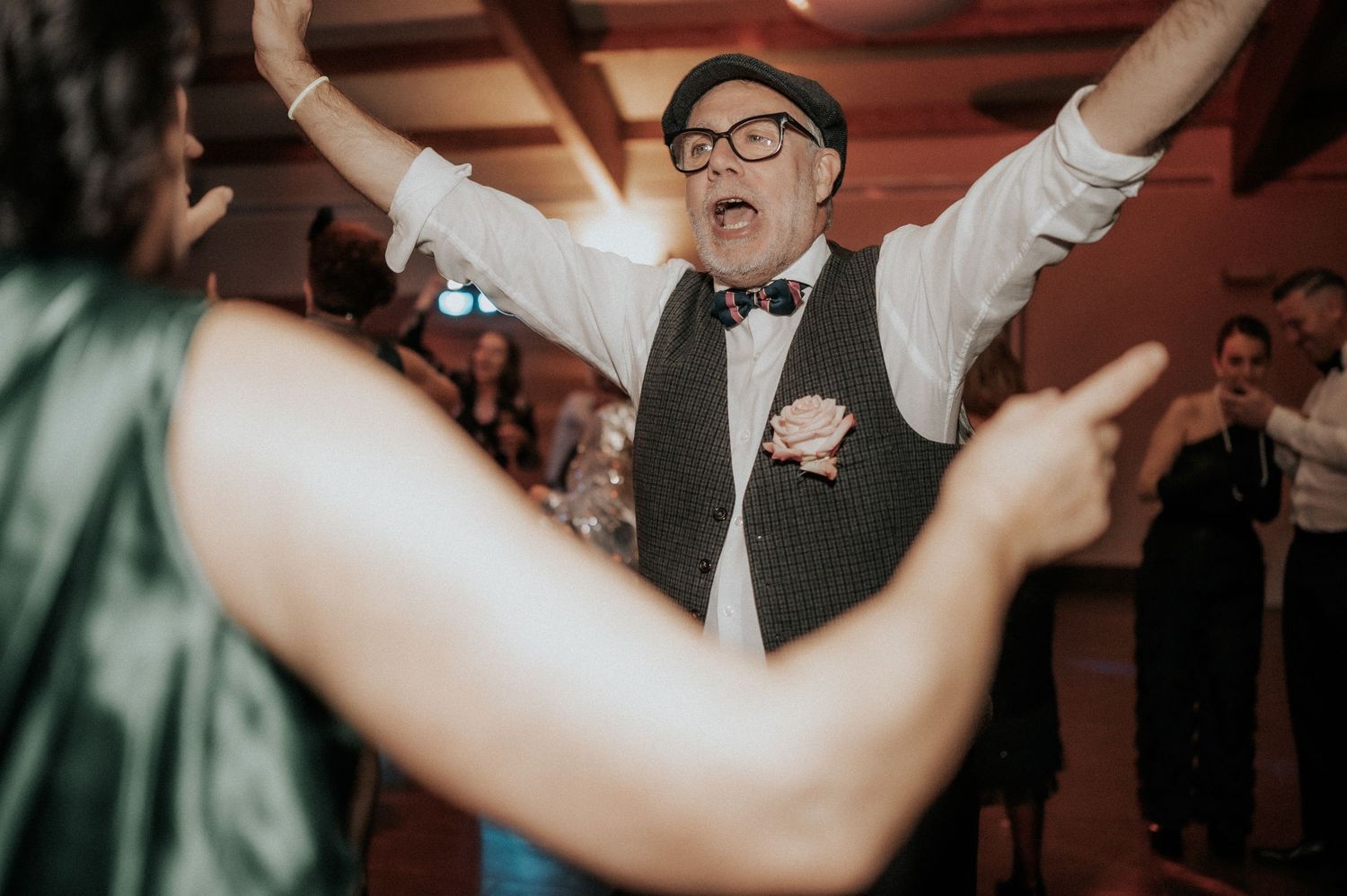 A person in a vest and glasses dances enthusiastically with raised arms at a lively indoor celebration.