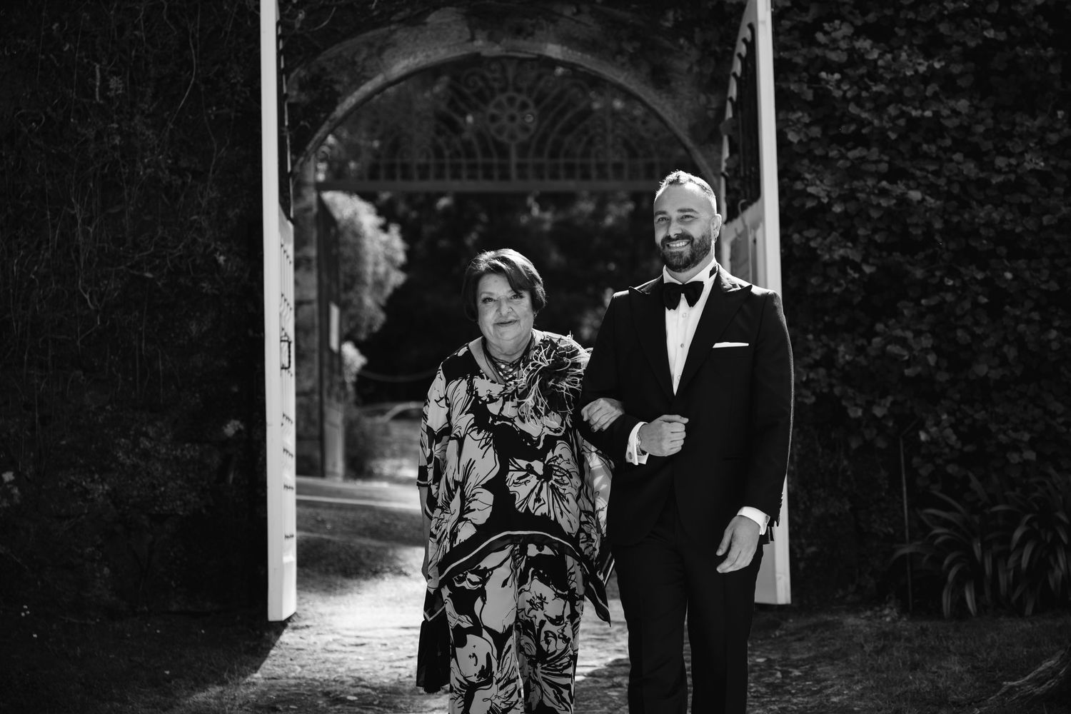 A couple walks together through a garden path surrounded by lush greenery and trees.