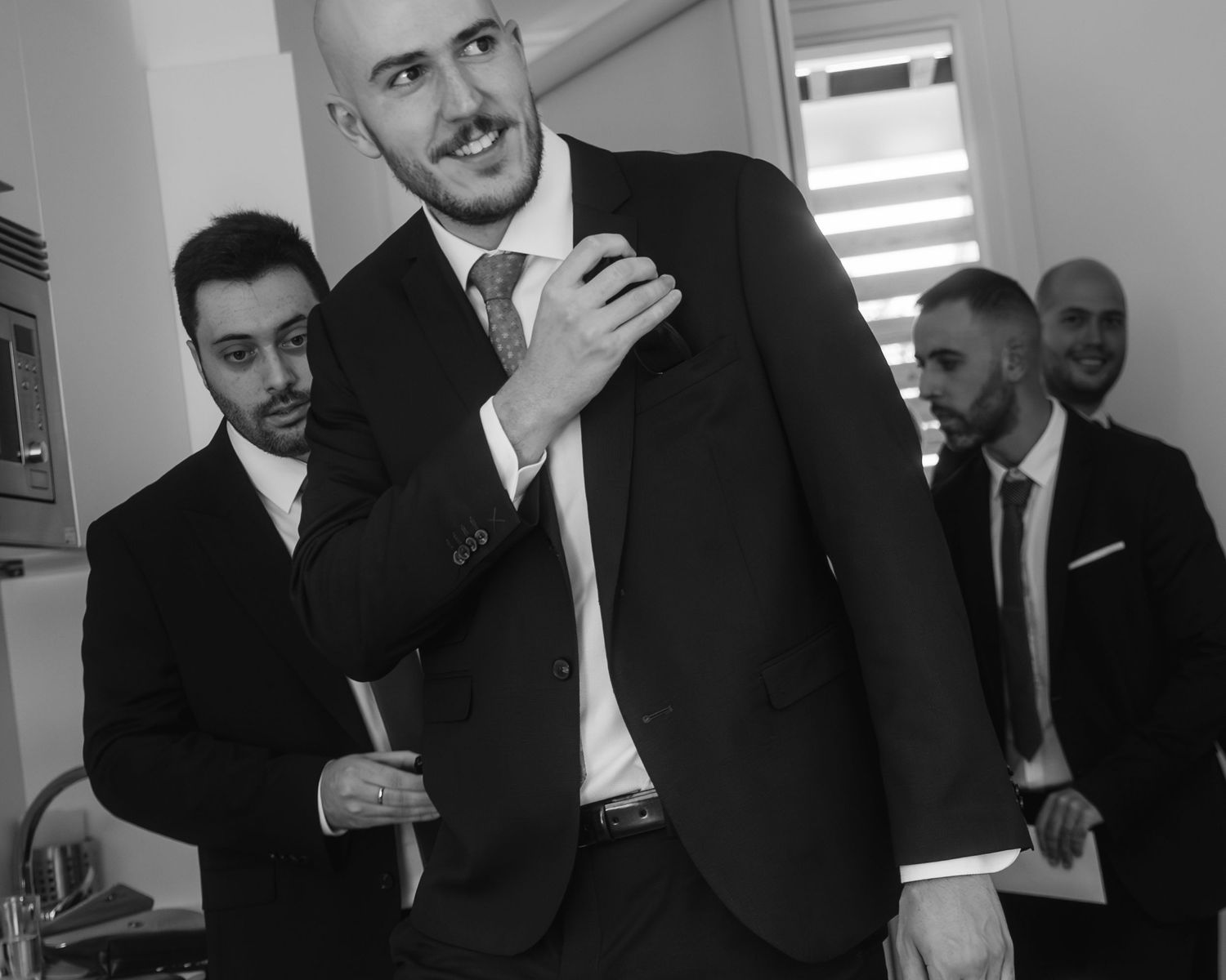 Black and white photo of groomsmen getting ready before a wedding ceremony in a bright room.