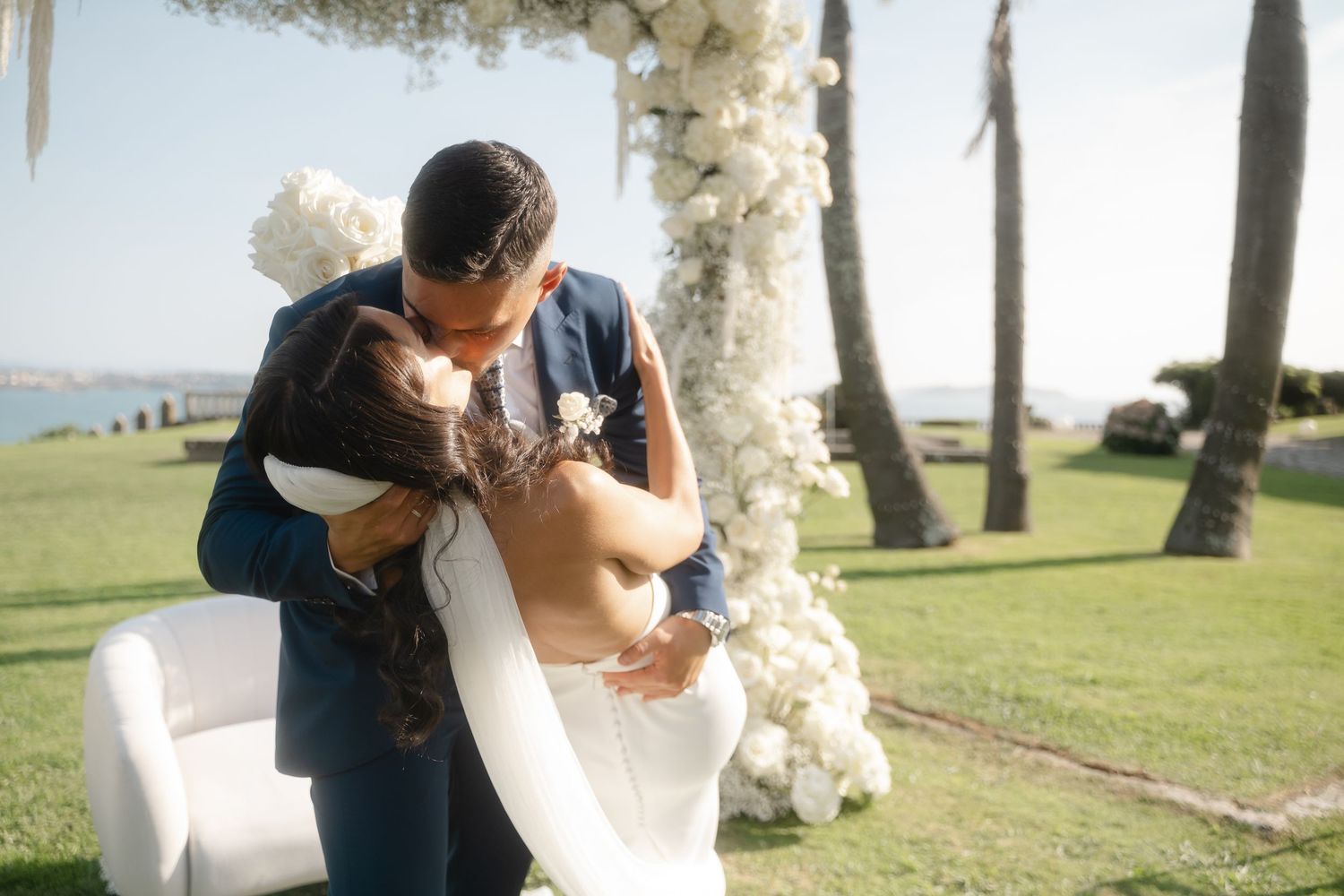 A couple in wedding attire shares an intimate dance together outdoors among tall trees on a sunny day.