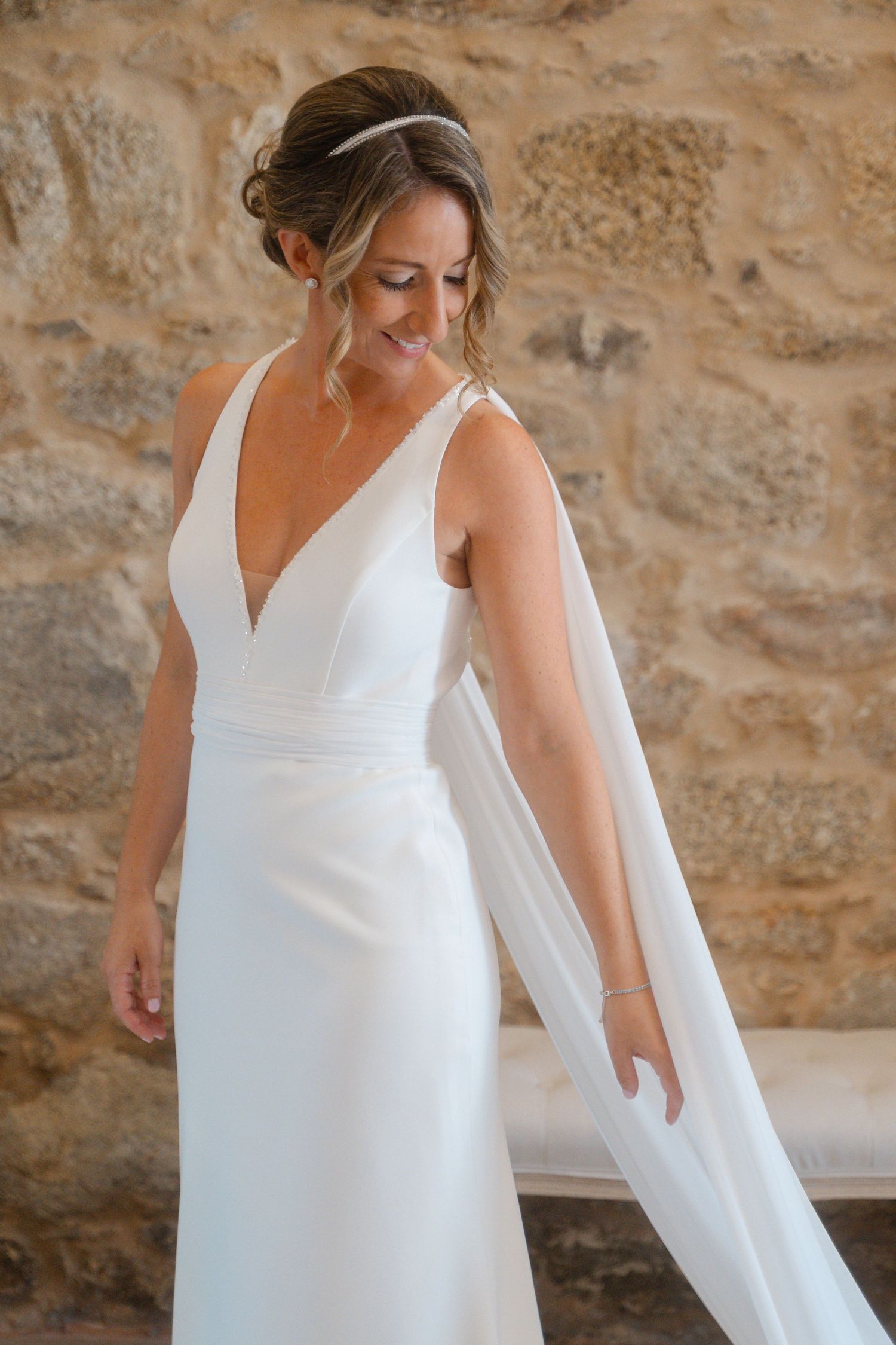 A person wearing a flowing white wedding dress stands against a rustic stone wall in elegant bridal attire.