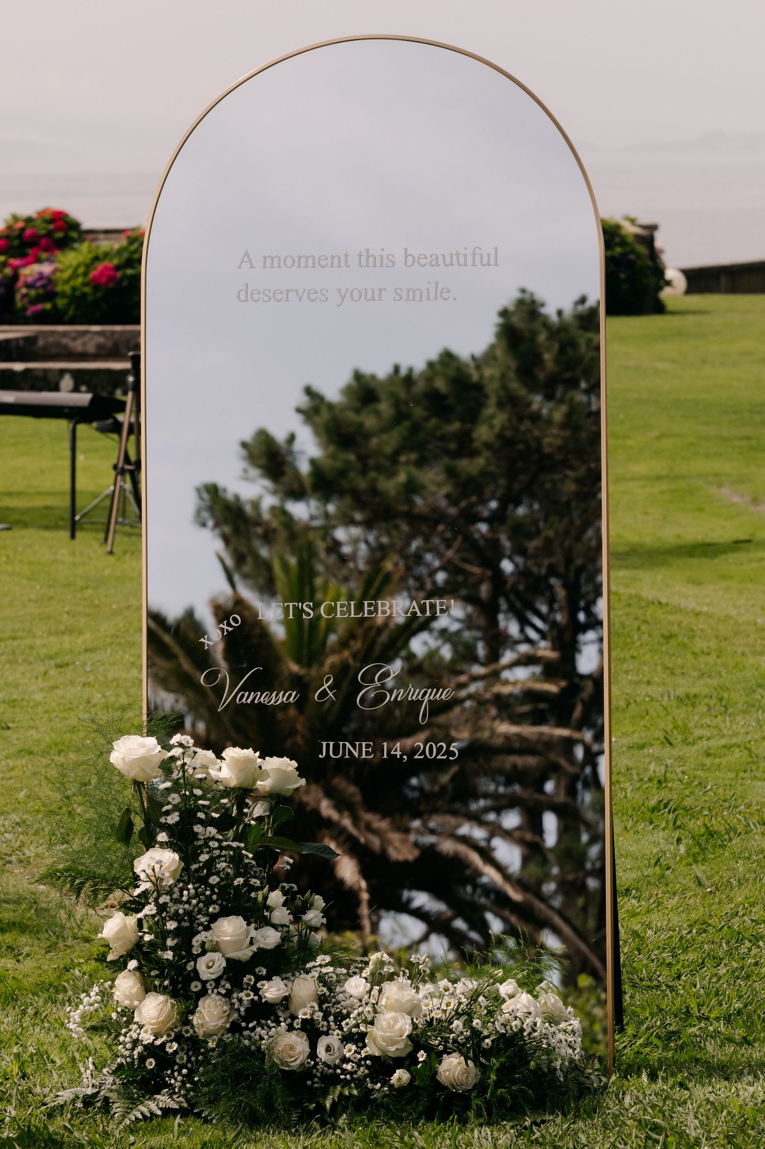 Clear acrylic wedding sign with white roses and greenery decorations on a lush green lawn at an outdoor ceremony.