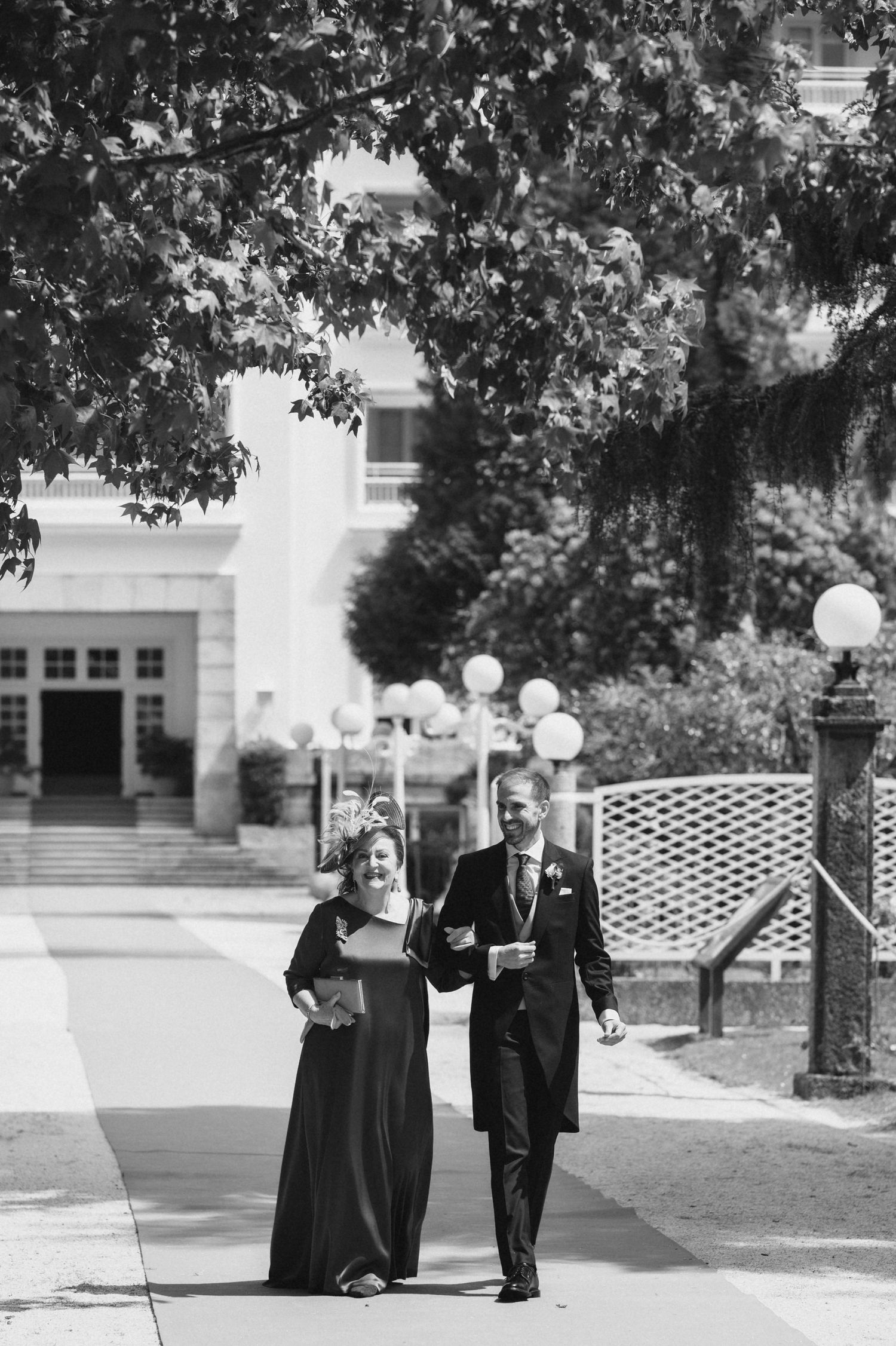 Couple strolls together through a park-like setting with trees and benches visible in the background.