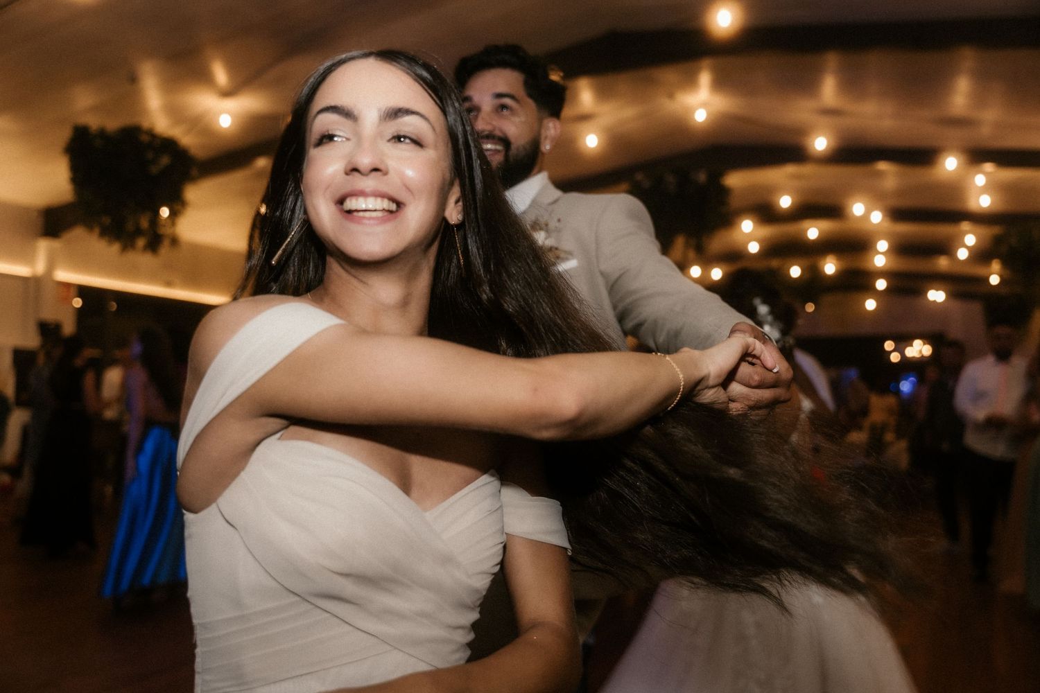 Wedding guests dance and celebrate under warm string lights at an indoor reception venue.