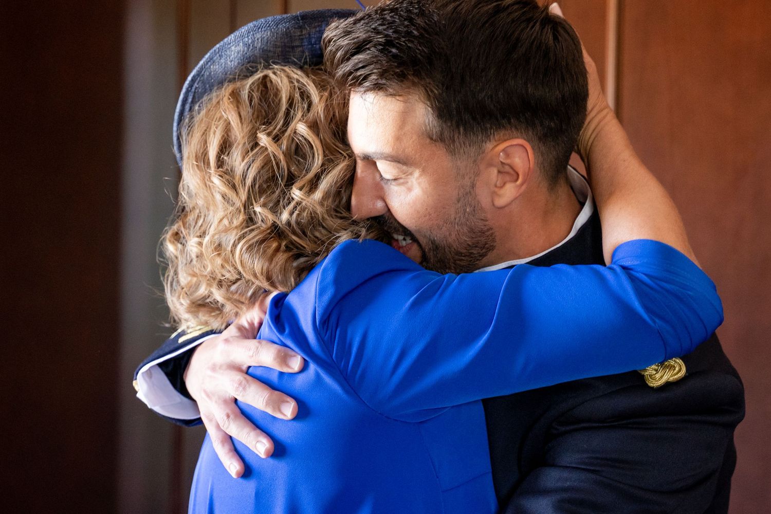 Two people share an emotional embrace, with one wearing a bright blue top and the other in a dark suit.
