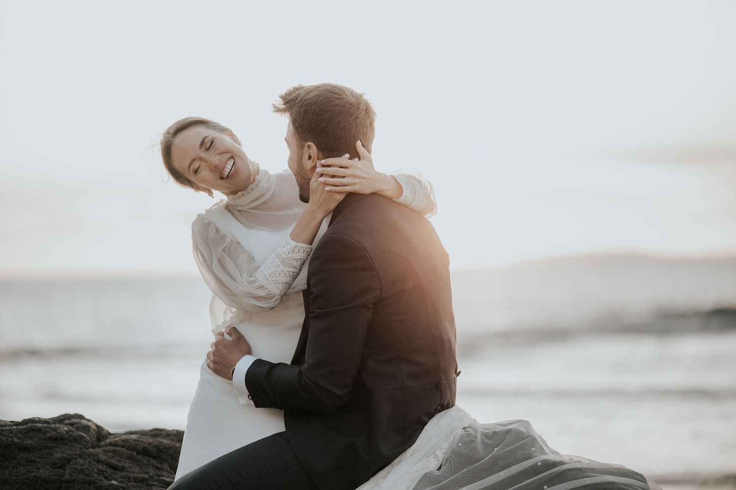 A romantic couple embraces on a rocky beach at sunset with ocean waves in the background.