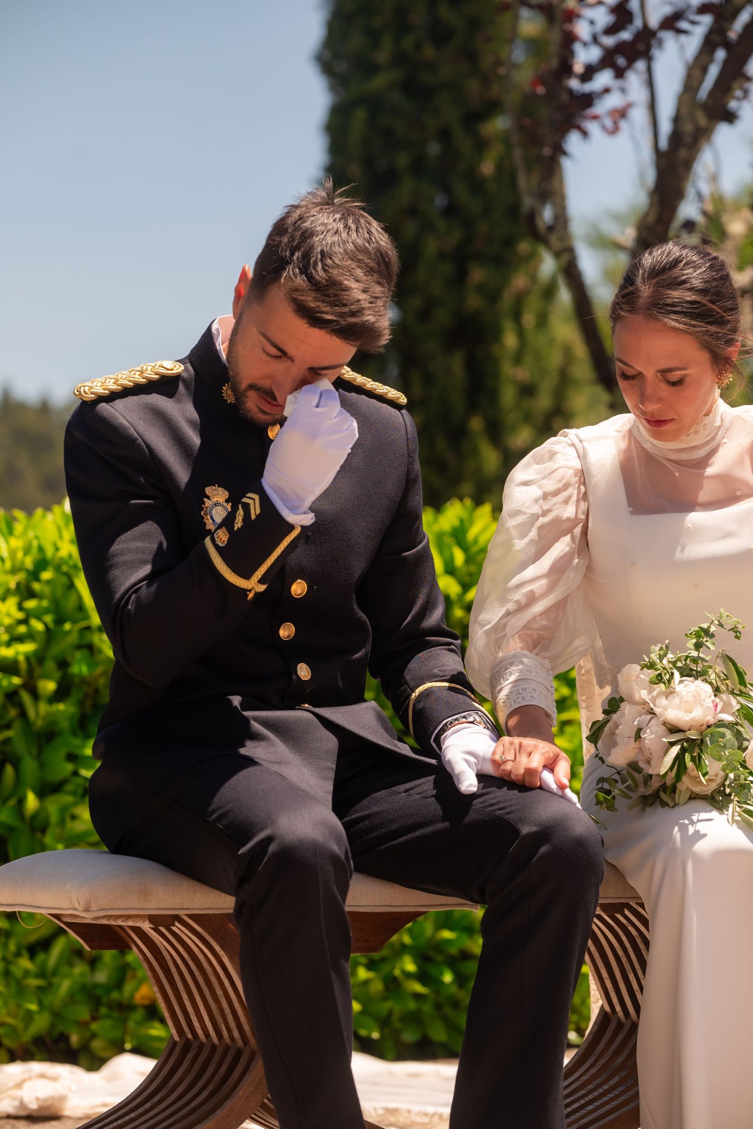 Military groom in dress uniform wipes tears during emotional outdoor wedding ceremony.