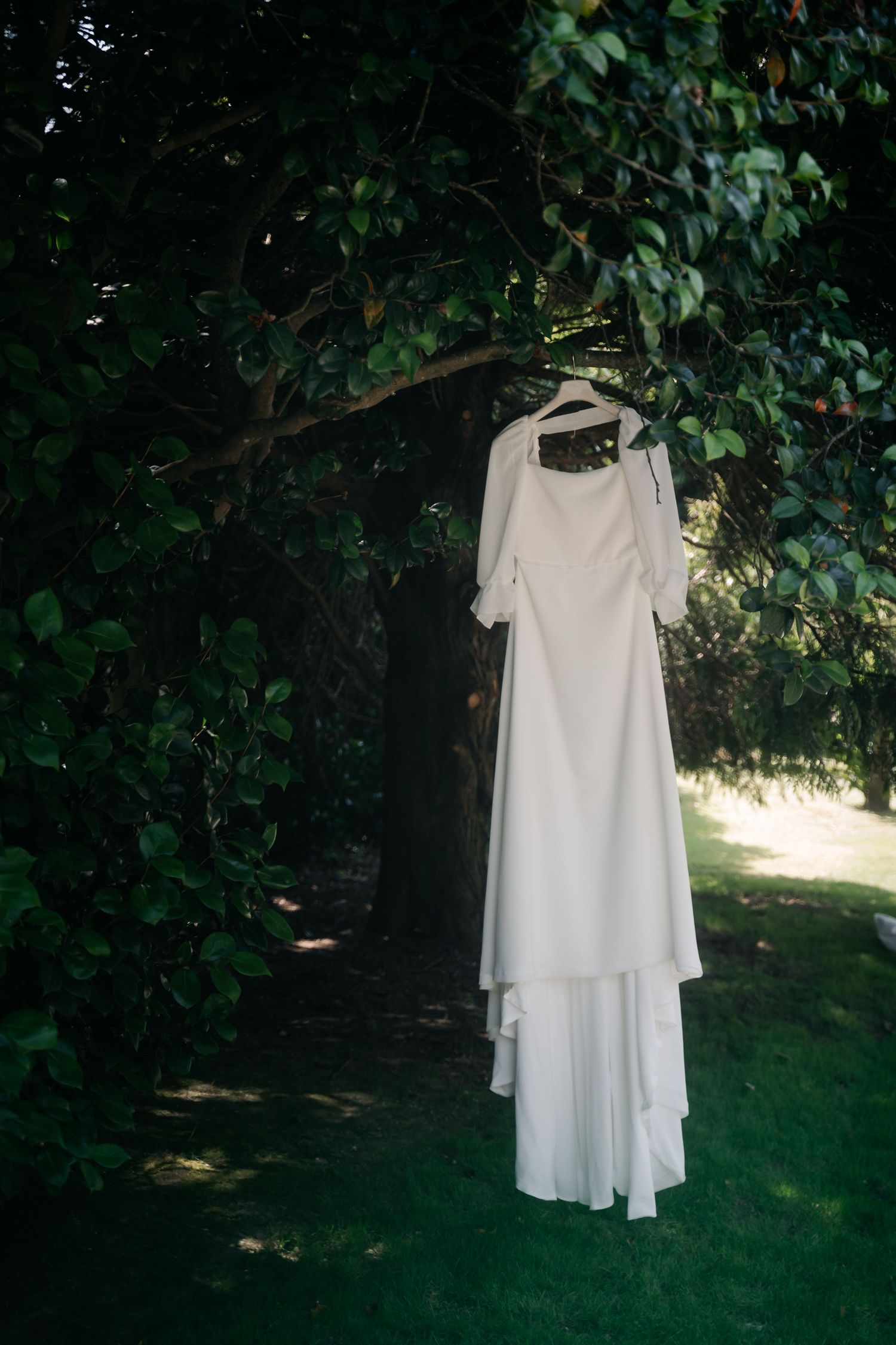 A white layered dress hangs on a wooden hanger against lush green foliage in natural outdoor lighting.