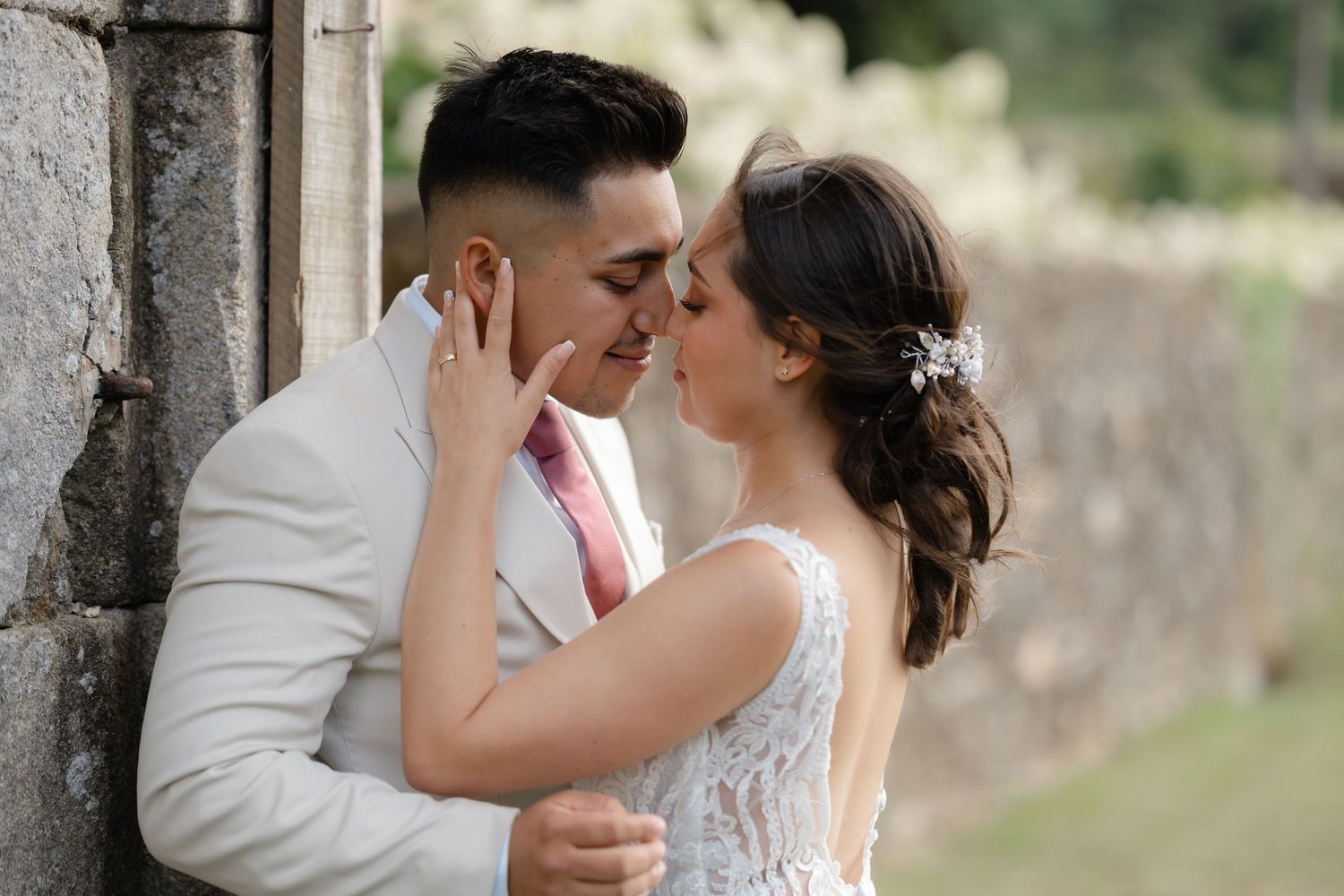 A romantic couple in wedding attire shares an intimate kiss against a stone wall backdrop in an outdoor setting.