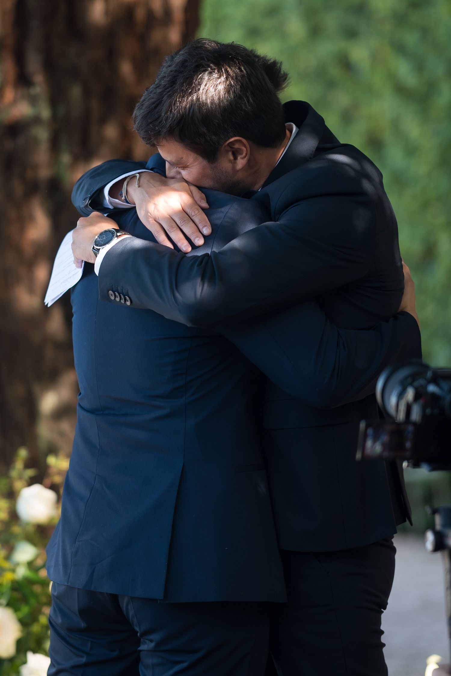Two people in dark formal attire share an emotional embrace outdoors against a green natural background.