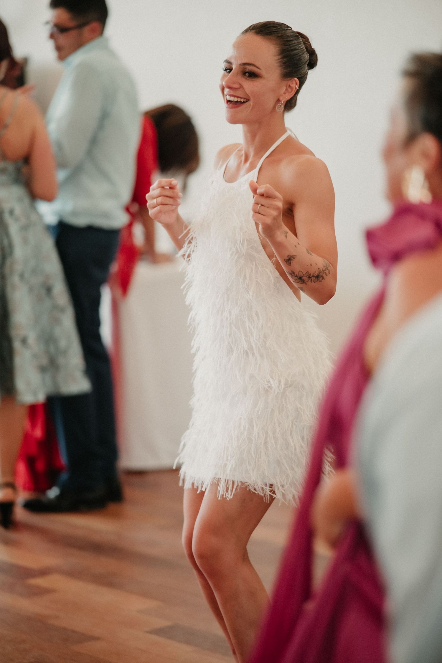 A bride in a feathered white dress dances and celebrates with guests at a wedding reception.