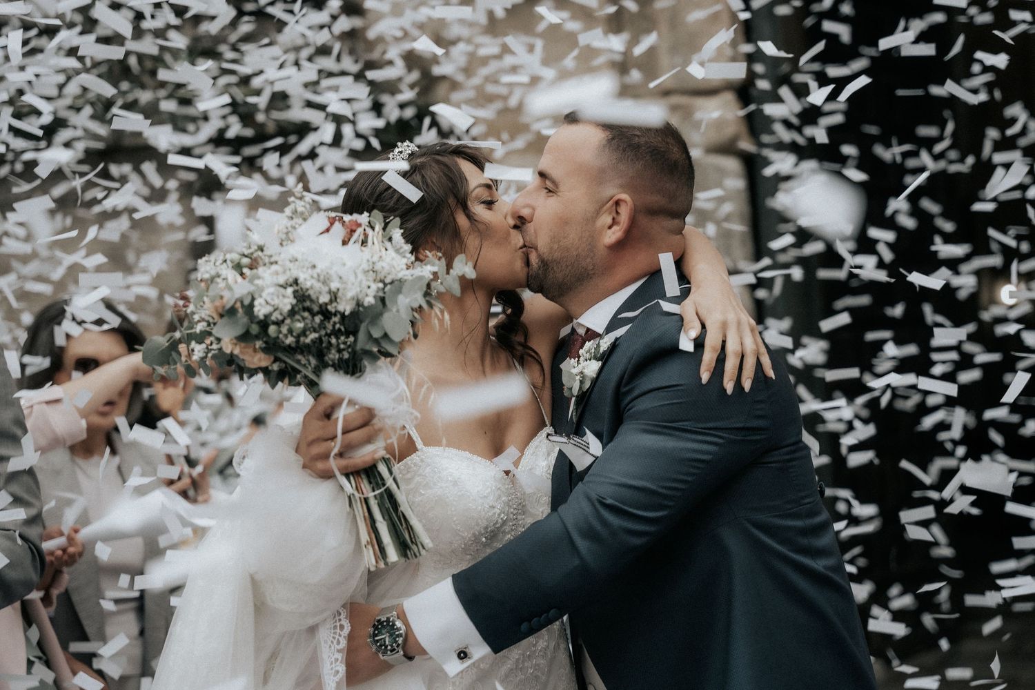 A joyful wedding couple shares a kiss amid a shower of white confetti while holding a beautiful floral bouquet.