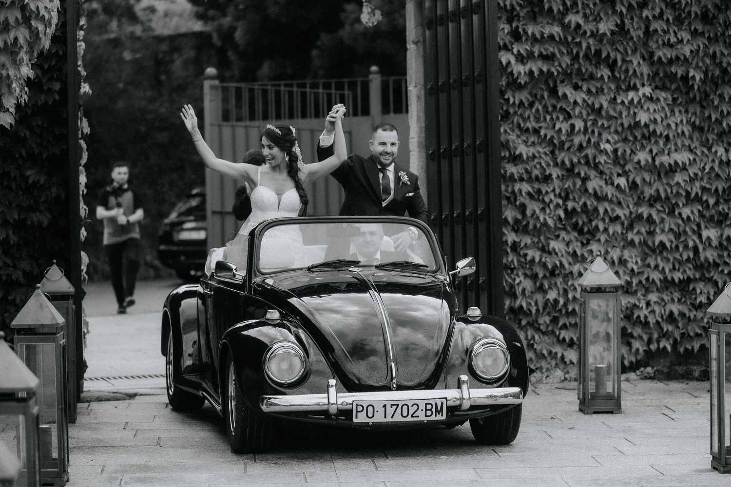 A vintage black Volkswagen Beetle convertible with a couple celebrating in formal attire on a city street.