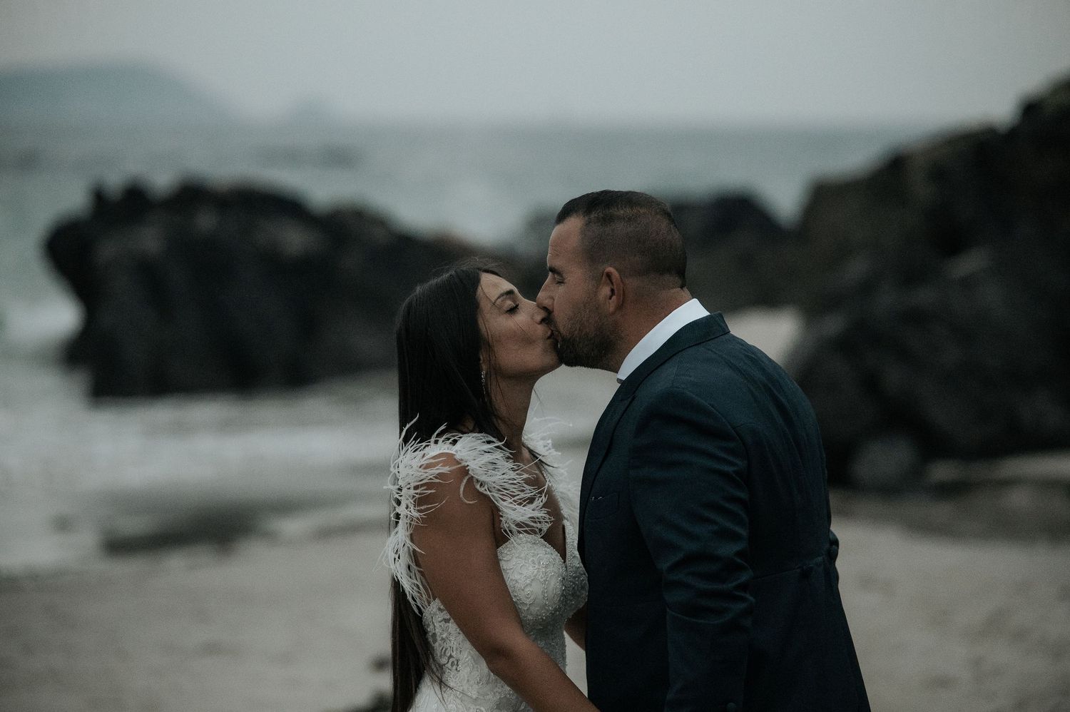 A couple in formal wedding attire share an intimate moment on a misty beach with rocky cliffs in the background.