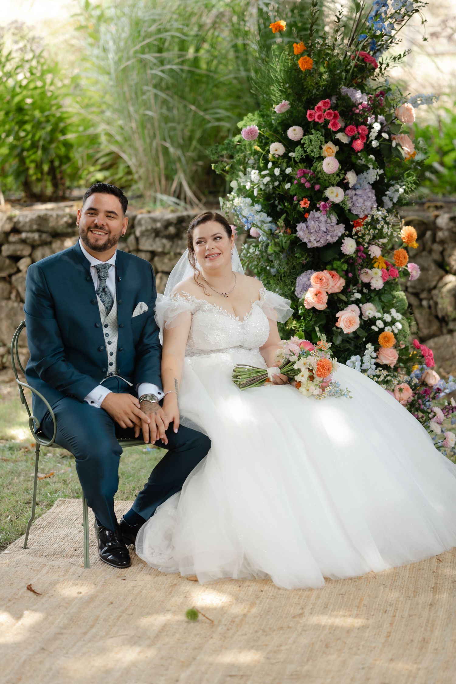 Wedding couple sitting together on stone steps surrounded by vibrant floral arrangements in a garden setting.