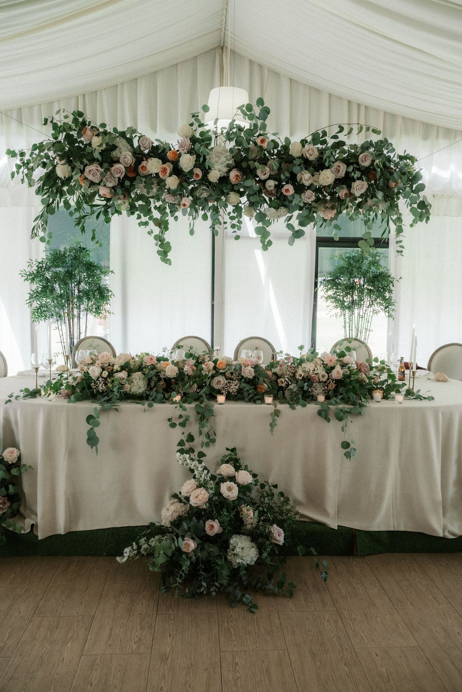 Elegant wedding reception setup with white draping and lush floral arrangements featuring greenery and blush roses.